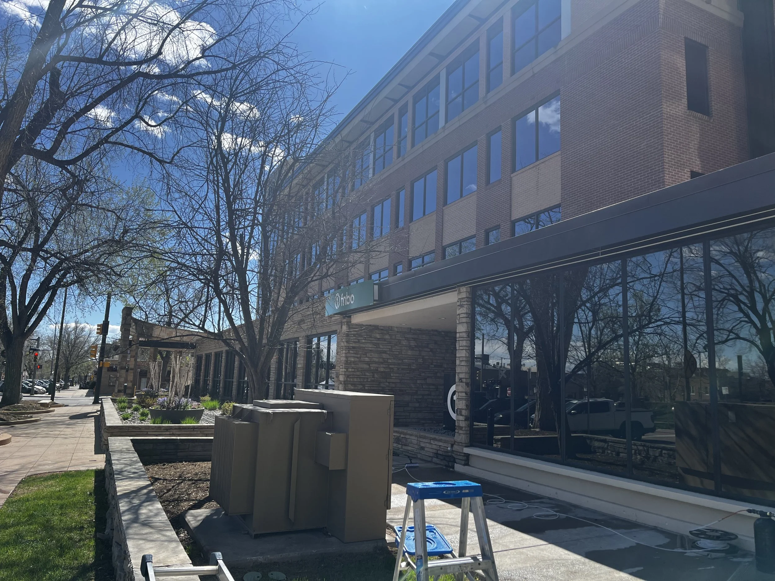 A multi-story brick and glass building under construction or renovation, with blue sky and leafless trees in front, and construction materials and equipment on the sidewalk.