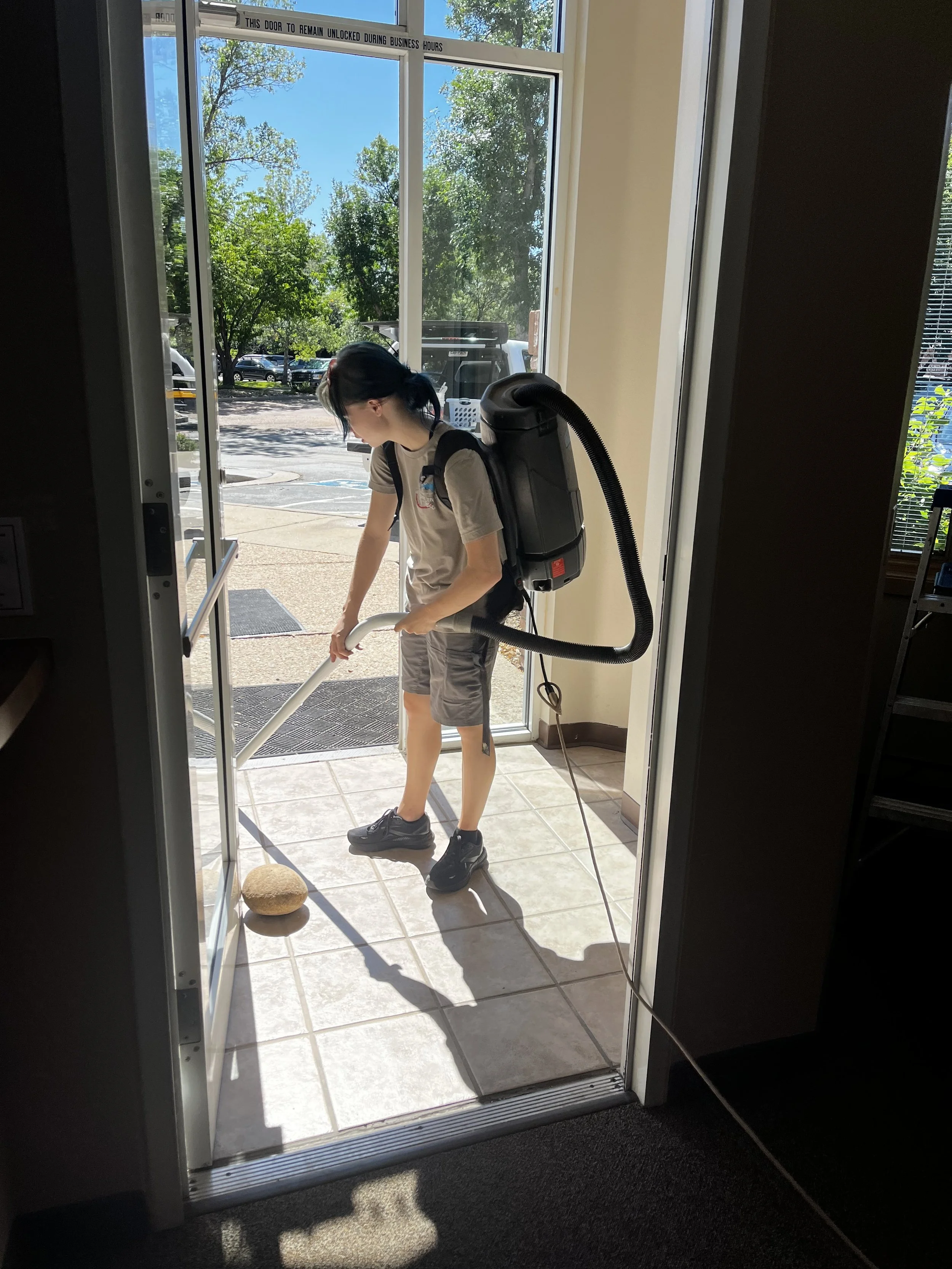 A person wearing a backpack vacuum cleaner is cleaning the entryway of a building, standing on a tiled floor near an open glass door revealing a sunny outdoor parking area with trees and parked cars.