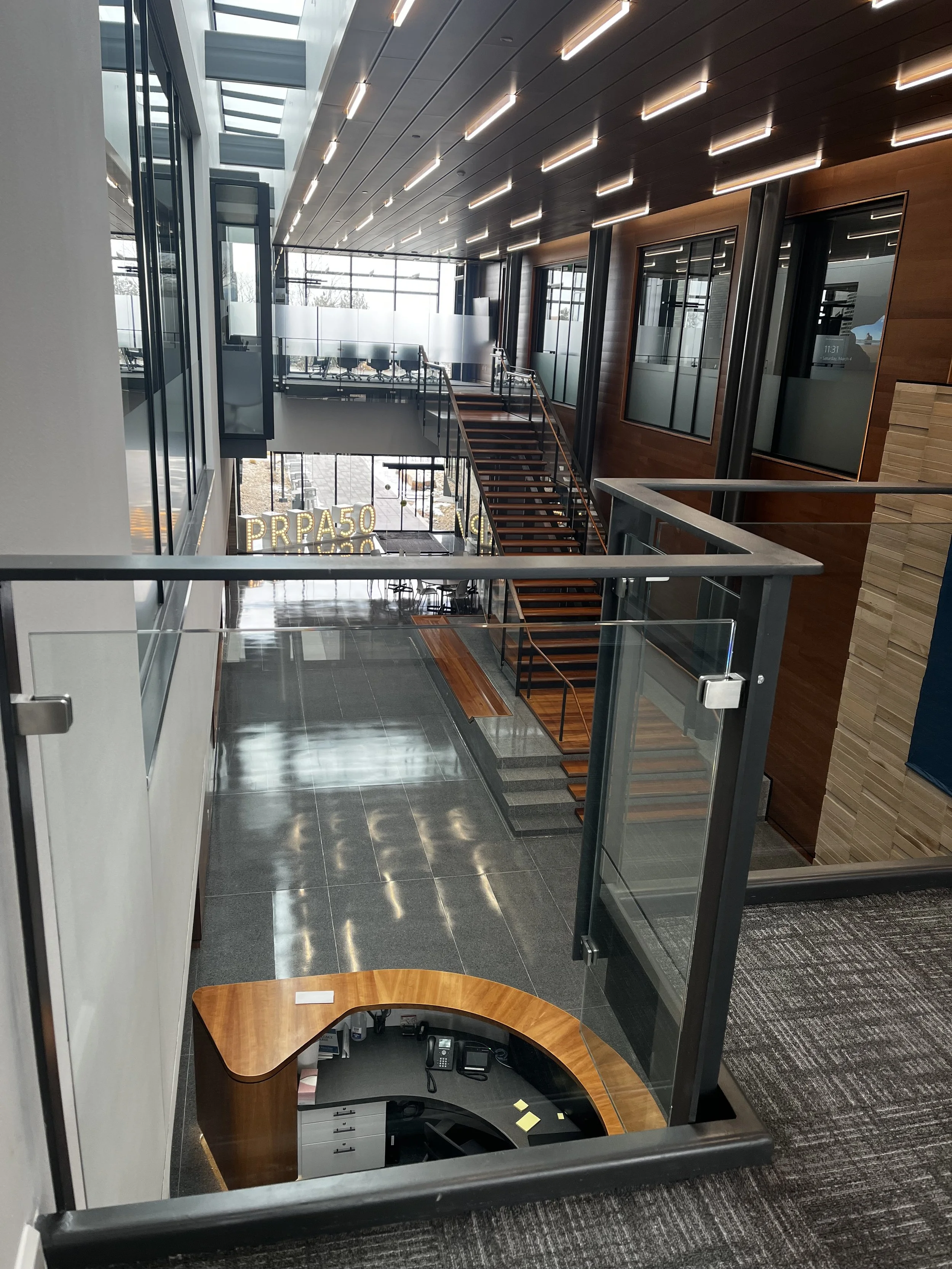 Interior of a modern office building lobby with a reception desk at the bottom, glass walls, and a staircase leading to an upper level. Large windows let in natural light, and there are wood panel accents on the walls and ceiling.