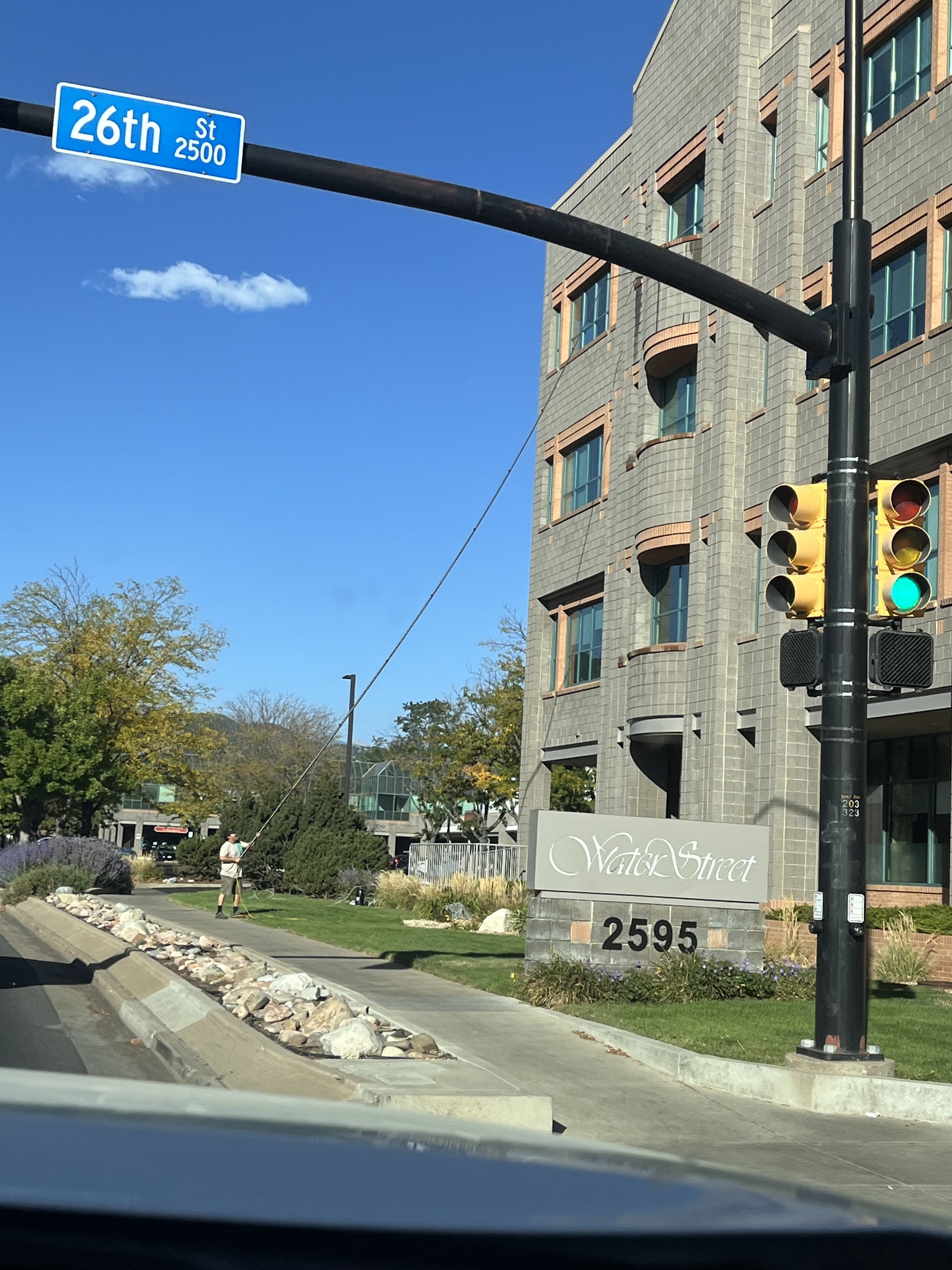 Street view showing a blue street sign for 26th Street, a traffic light with green signal, and a man watering plants in front of a modern building with a sign that reads 'Water Street' and the number 2595.
