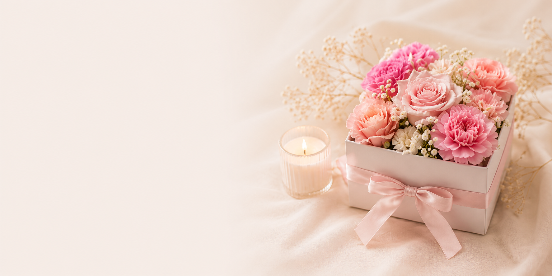 Pink and white floral arrangement in a white box with a pink ribbon, accompanied by a lit candle in a glass holder, on a soft surface with a light background.
