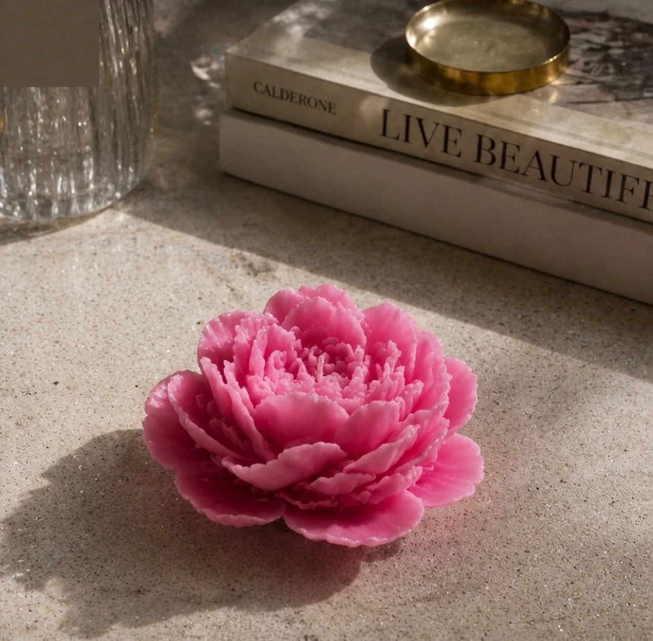 A pink artificial flower with layered petals sits on a beige textured surface. In the background, a clear glass container, a stack of books, and a brass tray are visible.