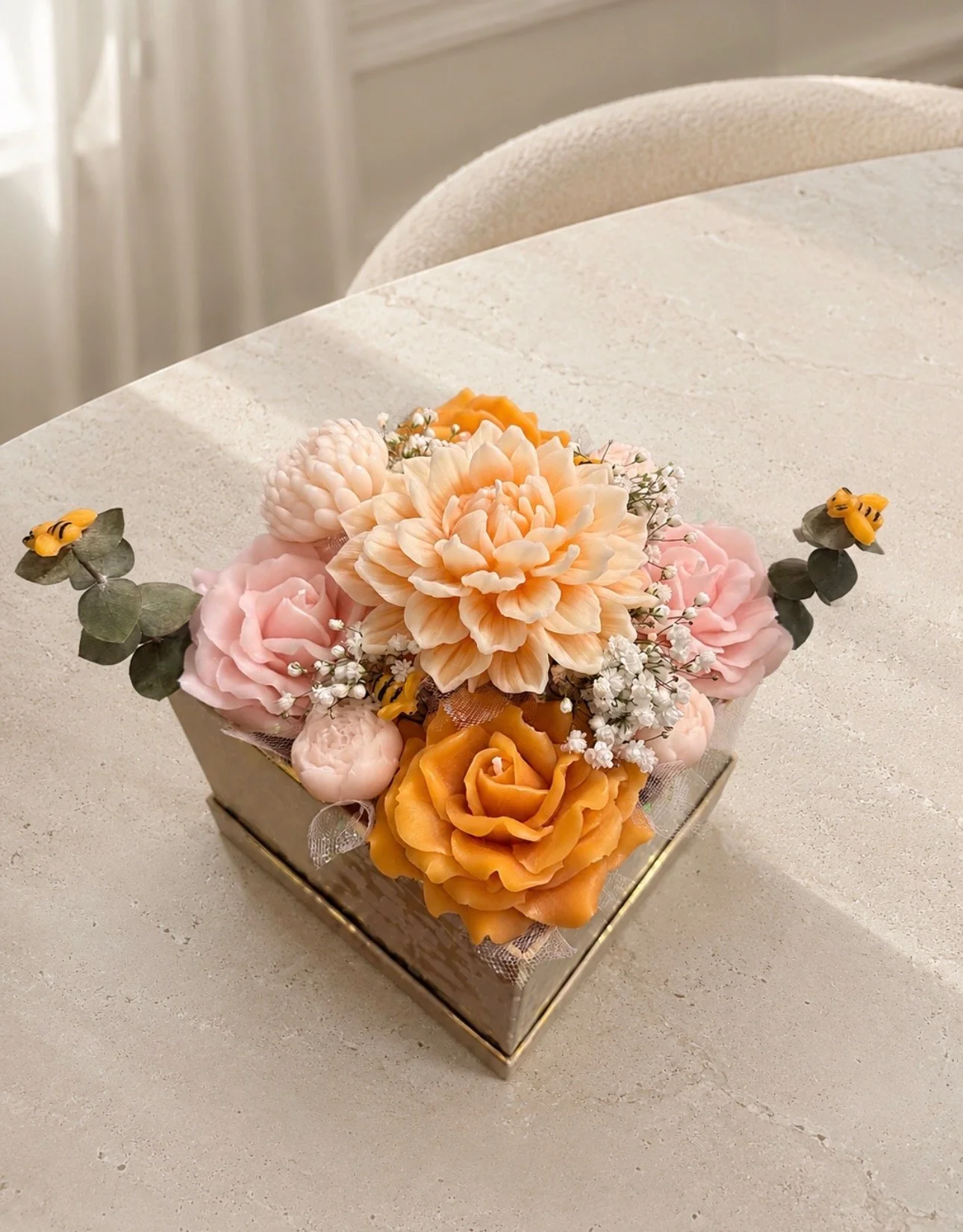 A floral arrangement in a square silver box with peach, pink, and white flowers, including roses and dahlias, accented with greenery and small white baby's breath, placed on a light-colored table.