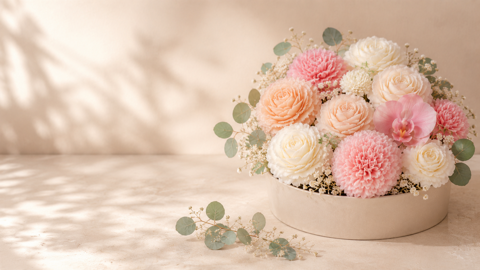 A round beige flower arrangement with pink, white, and cream roses, add in pink and white cockscomb flowers, accented with small white baby's breath flowers and eucalyptus leaves, set against a neutral background on a beige surface.