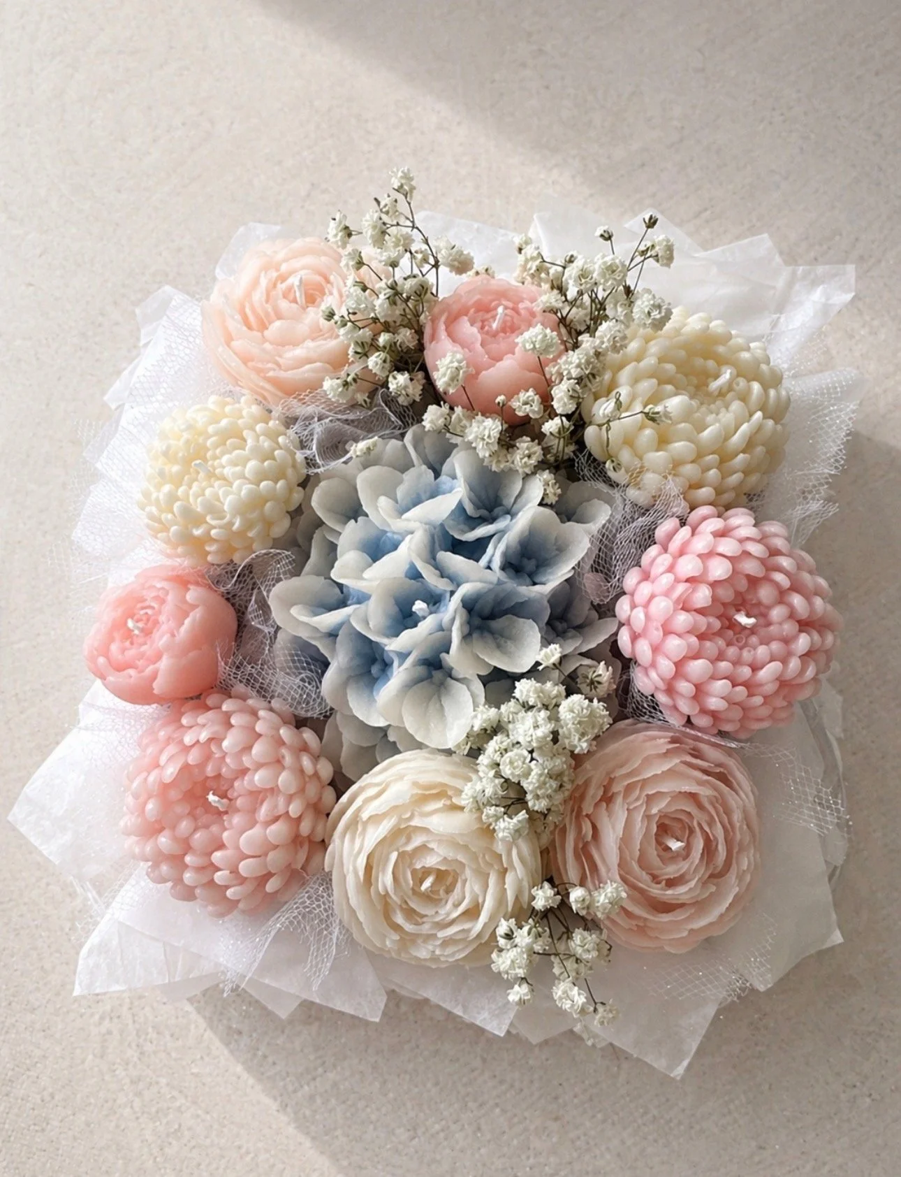 Arrangement of decorative candles shaped like flowers in pastel pink, blue, and cream colors, accented with white baby's breath flowers, wrapped in white mesh fabric.