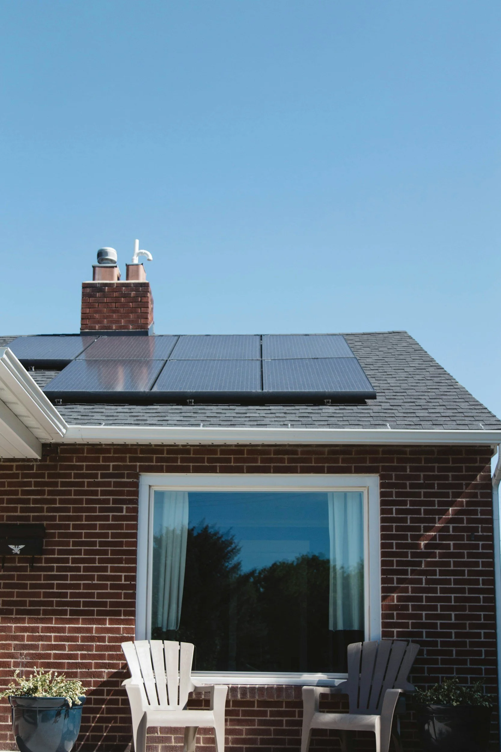 The image shows the exterior of a brick house with solar panels on the roof and a large window with white curtains. There are two white chairs in front of the window, with a blue potted plant next to the chairs and another pot on the right side. The 