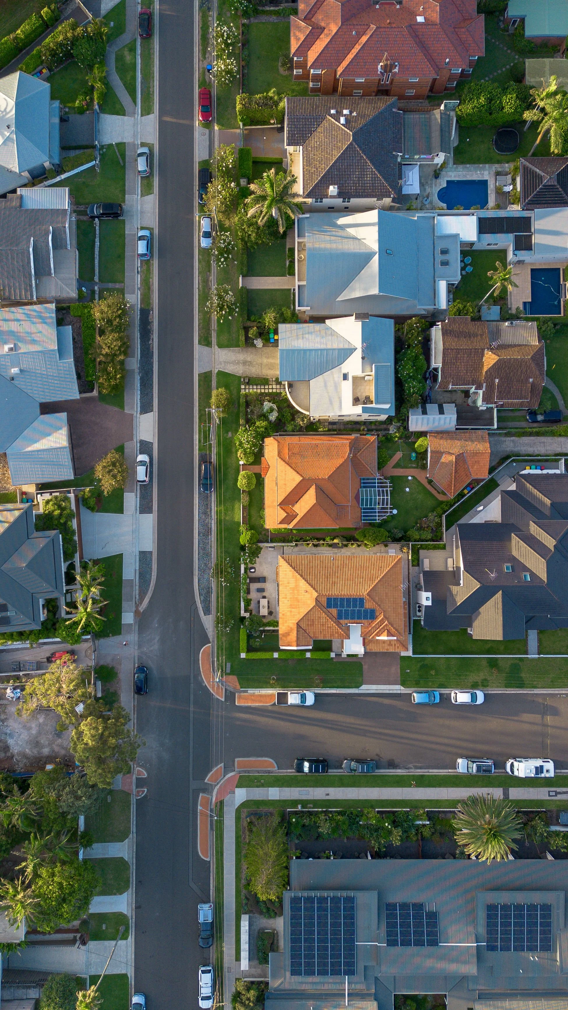 An aerial view of a residential neighborhood showing houses with tiled roofs, some with solar panels, manicured lawns, street trees, parked cars along the curb, and sidewalks.