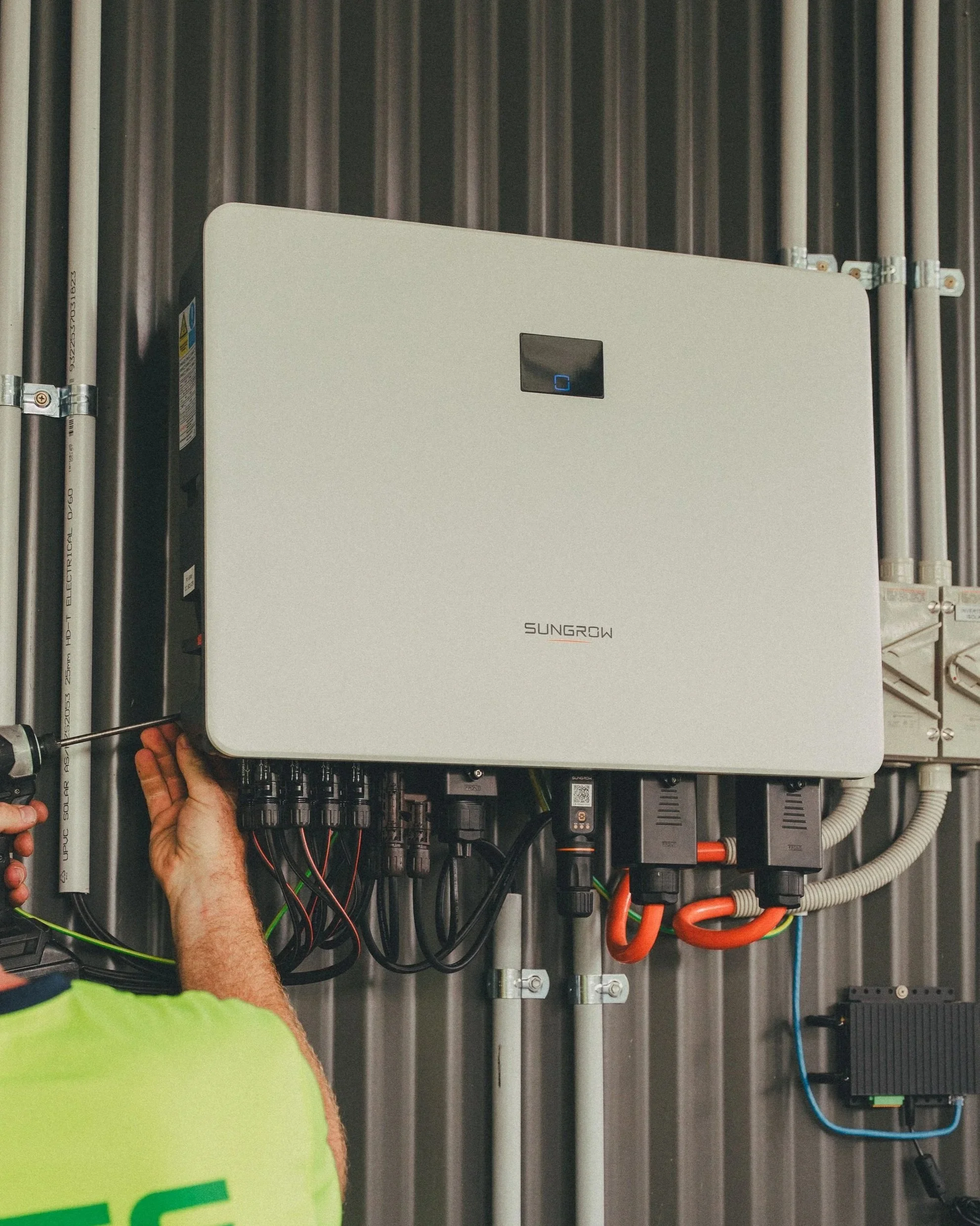 An individual wearing a yellow safety vest installs or maintains an electrical solar panel inverter system on a metal electrical panel board, with various wires and connections visible.
