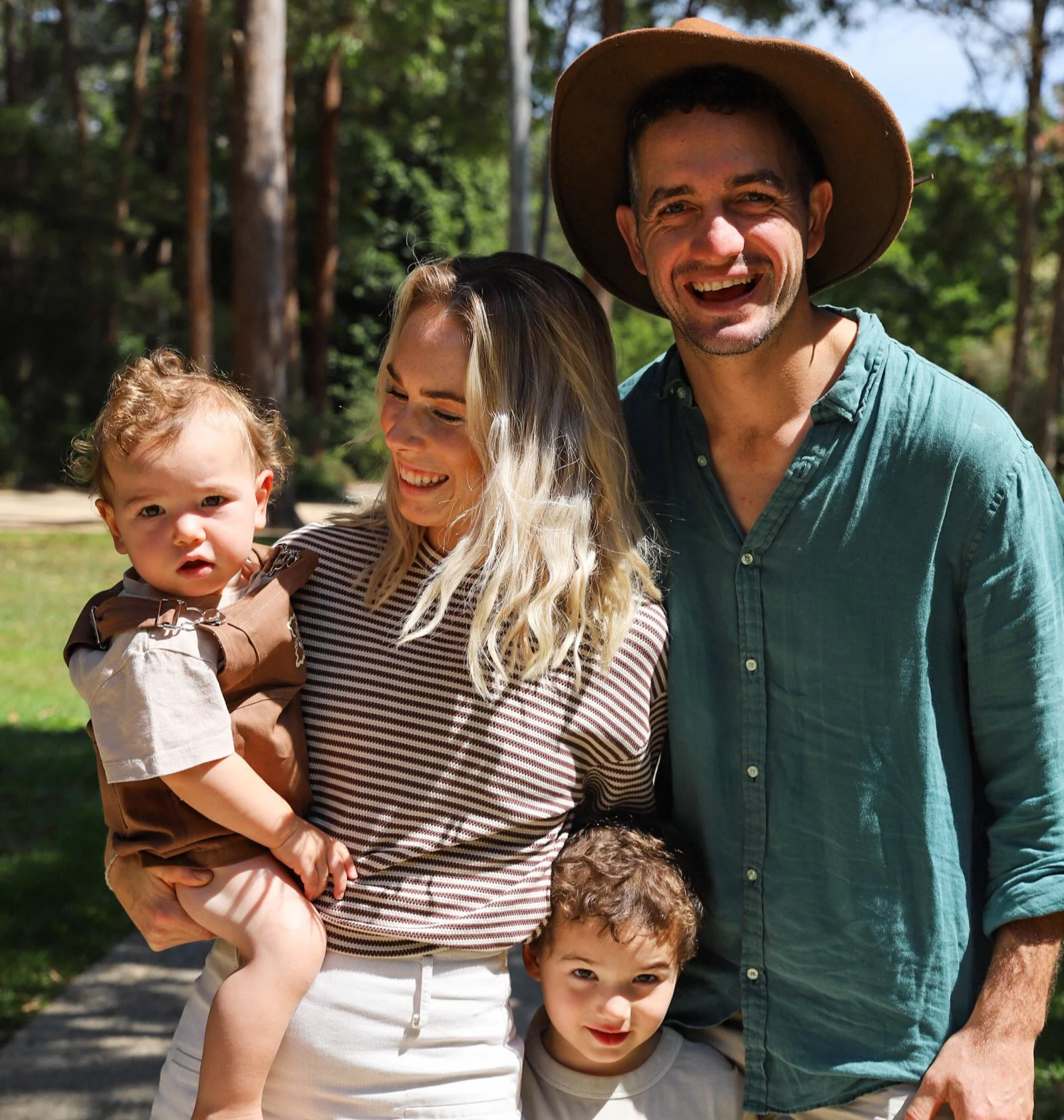 A happy family outdoors, including a man with a wide-brimmed hat, a woman holding a small child, and two young boys, all smiling and enjoying a sunny day in a park with trees in the background.
