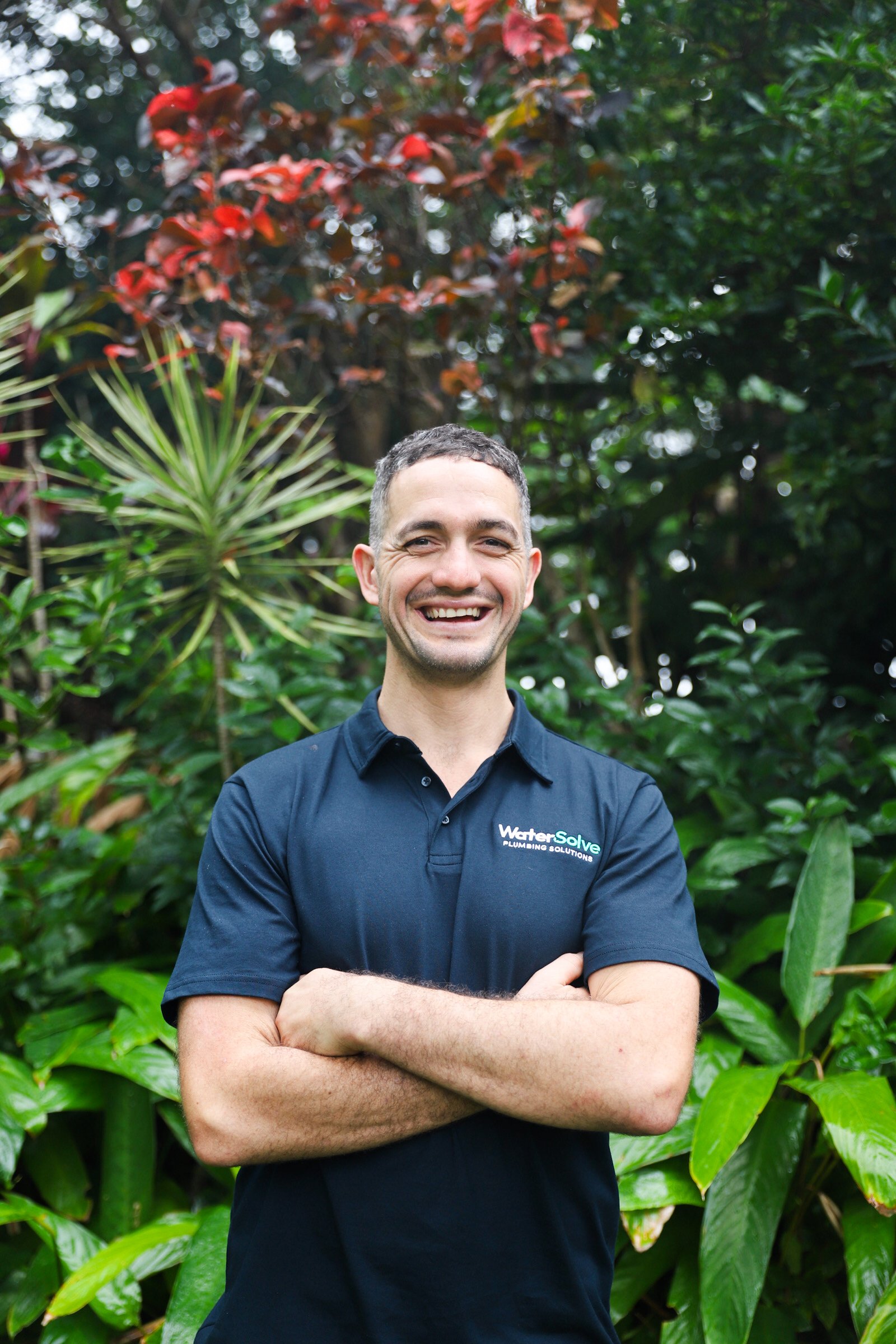 A smiling man with short dark curly hair, wearing a dark blue polo shirt with a logo that says 'WaterSolve Plumbing Solutions', stands with arms crossed in front of lush green plants and trees.