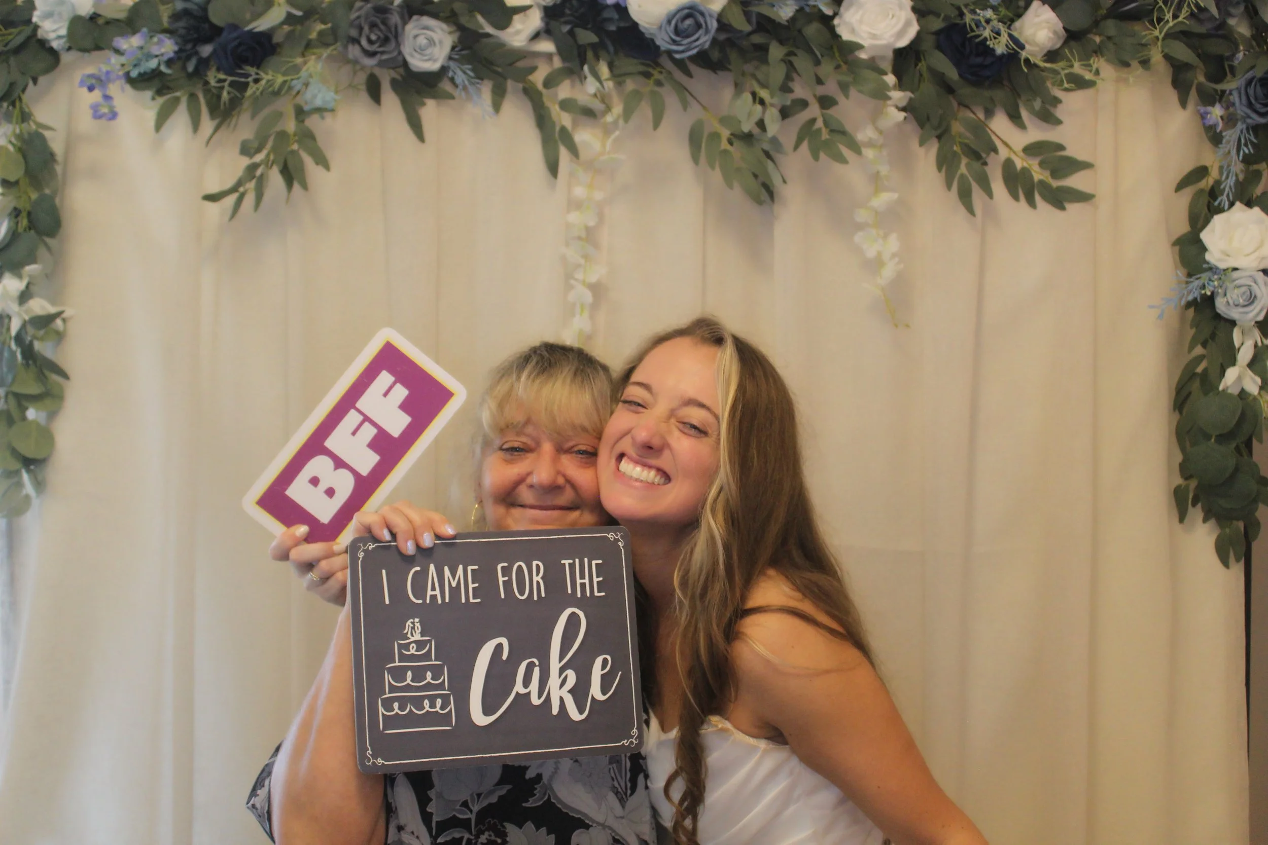 Two women smiling, one holding a sign that reads 'I came for the cake' and the other holding a birthday sign, standing in front of a floral backdrop.