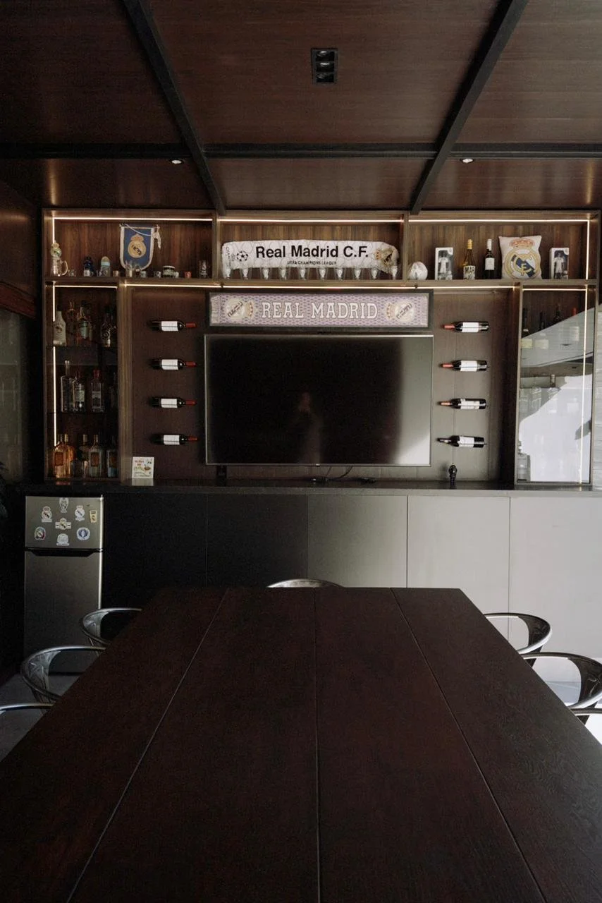 Una sala de reuniones con una mesa de madera oscura, en el fondo una pared de madera con estanterías que albergan botellas, objetos decorativos, y mantas con los lemas del Real Madrid.