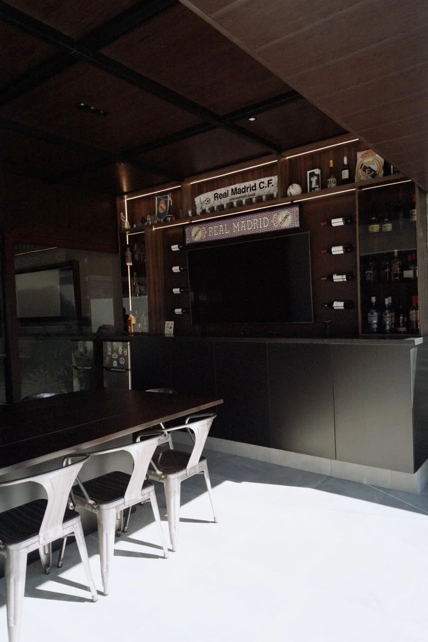 Bar en una casa con decoración del Real Madrid, incluyendo una barra negra, estanterías con botellas de alcohol y una televisión.