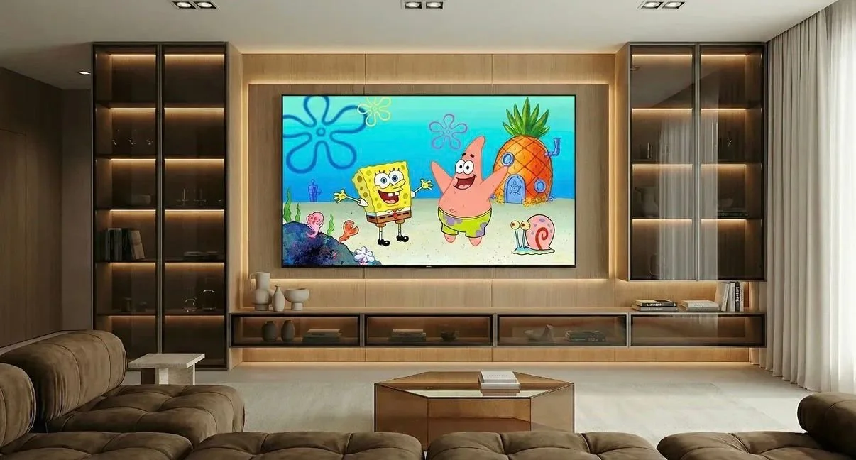 Salón moderno con un televisor grande que muestra a personajes de dibujos animados, SpongeBob y Patricio, en una escena submarina. La sala tiene sofás de colores neutros, una mesa de centro de madera, y estanterías con libros y decoraciones.