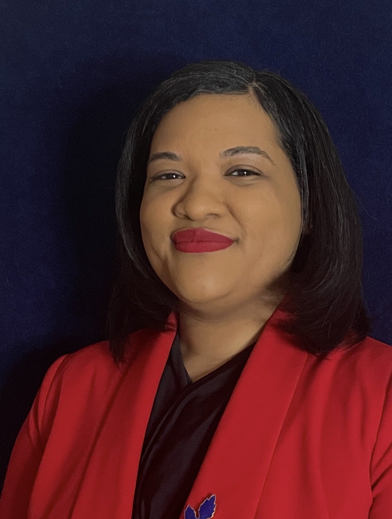 A woman with shoulder-length black hair, wearing red lipstick, a red jacket, and a black top, standing in front of a dark blue background.