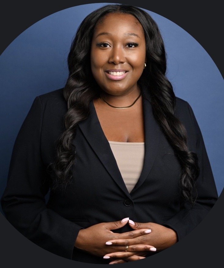 Professional portrait of a smiling African American woman with long wavy hair, wearing a black blazer and beige top, standing against a blue and dark background.