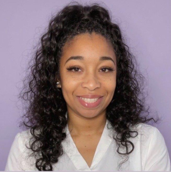 Portrait of a young woman with curly hair, wearing a white top, smiling, against a purple background.