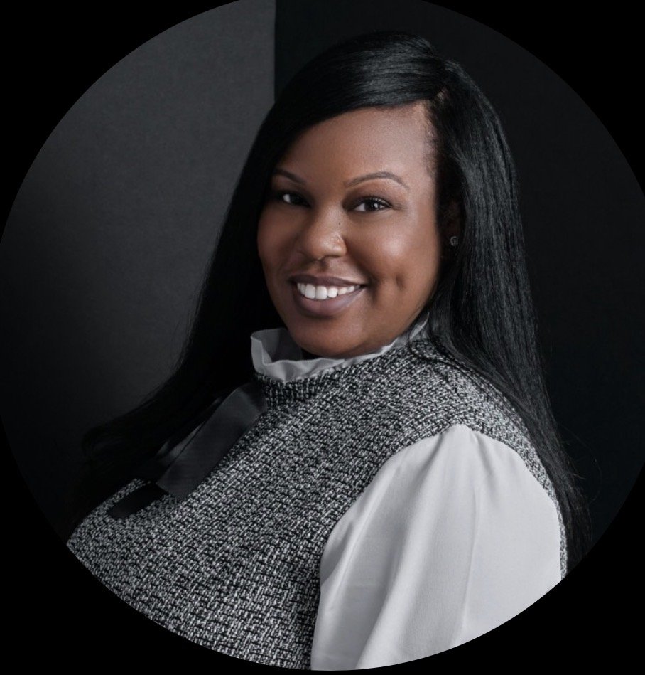 A smiling African American woman with long black hair, wearing a white blouse and a textured black and white vest, against a dark background.