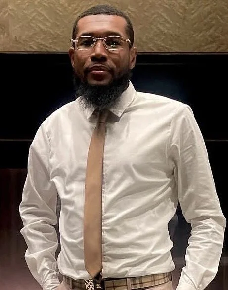 A man with glasses, a beard, wearing a white shirt, beige tie, and plaid pants, standing indoors against a dark background with a beige textured wall.