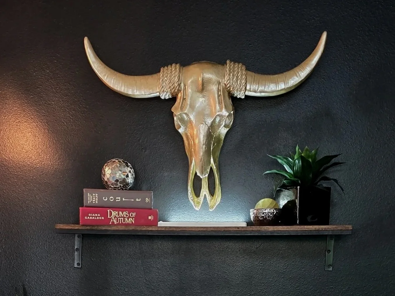Decorative wall sculpture of a buffalo skull mounted on a black wall above a wooden shelf with books, a small plant, and decorative objects.