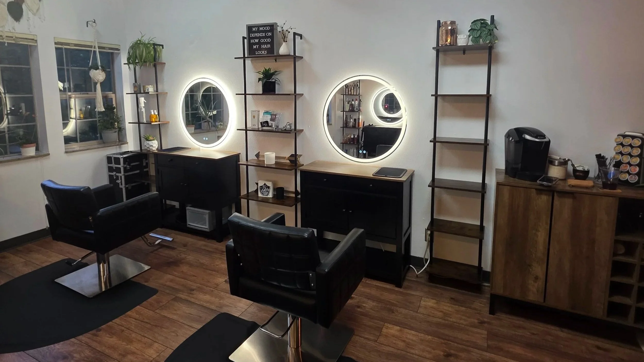 A salon or beauty studio with two illuminated round mirrors on the wall, shelves with decor items and plants, black chairs, and a coffee station.