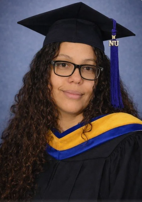 Woman wearing a black graduation cap and gown with yellow and blue accents, glasses, and long curly hair.