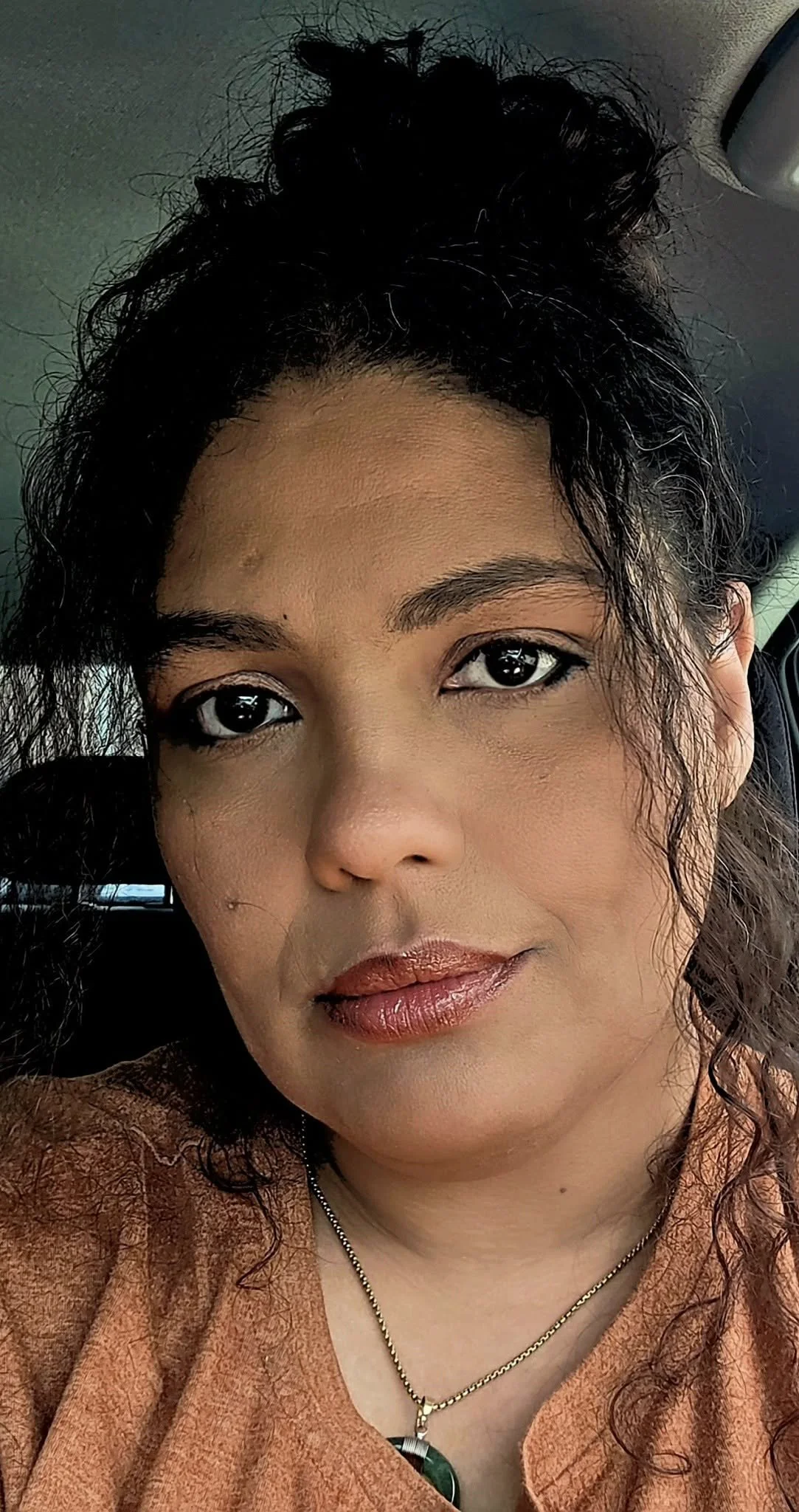 A woman with curly dark hair styled in a high bun, wearing makeup, a brown top, and a gold necklace with a green pendant, sitting inside a vehicle.