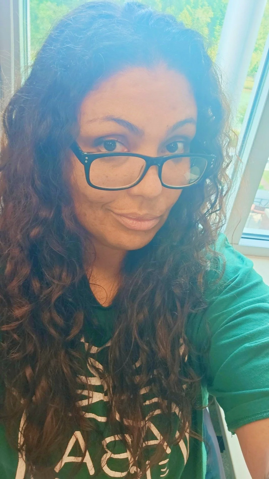 A woman with curly brown hair and glasses taking a selfie near a window, with green scenery in the background.
