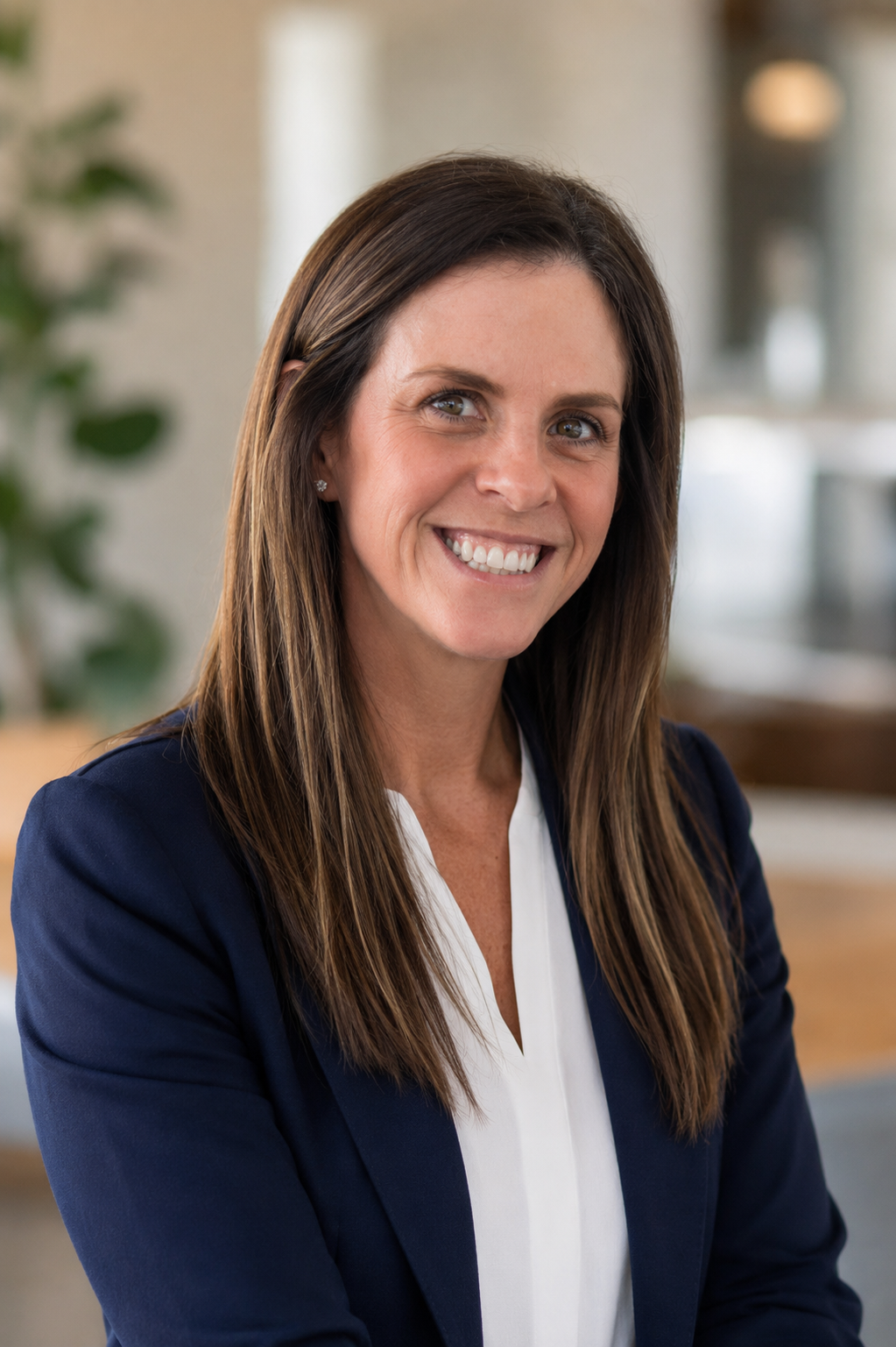 A smiling woman with long brown hair, wearing a white blouse and navy blazer, sitting in a bright, modern office environment.