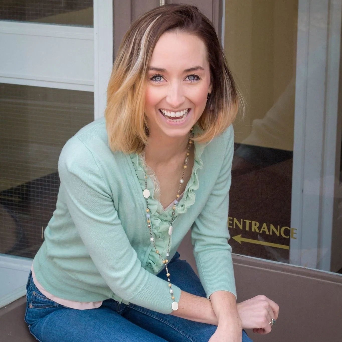 A woman with shoulder-length blonde hair smiling, wearing a light green sweater and a necklace, sitting outside near a glass door with a sign that says 'ENTRANCE' with arrows.
