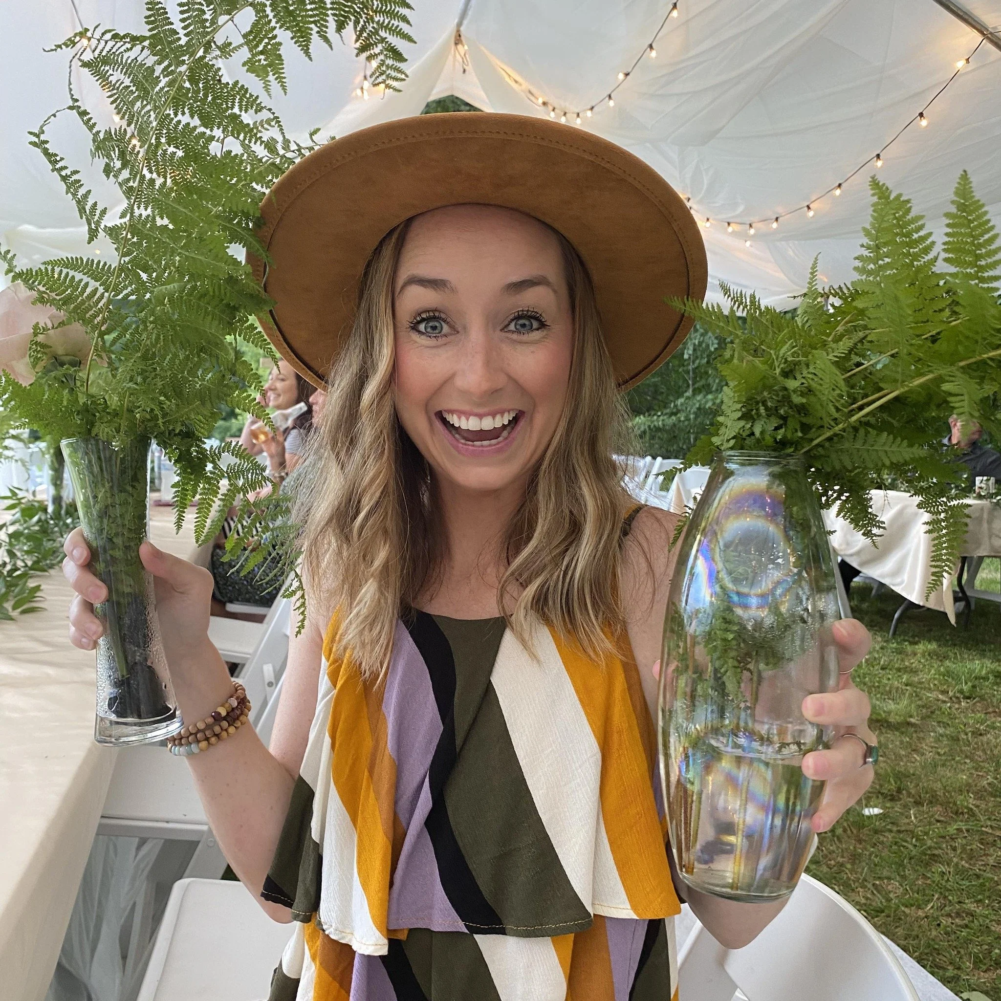 A woman with blonde hair and blue eyes, smiling widely, wearing a large brown hat and a multicolored scarf. She is holding a clear glass vase with ferns inside. There are string lights and a white drape in the background at an outdoor event.