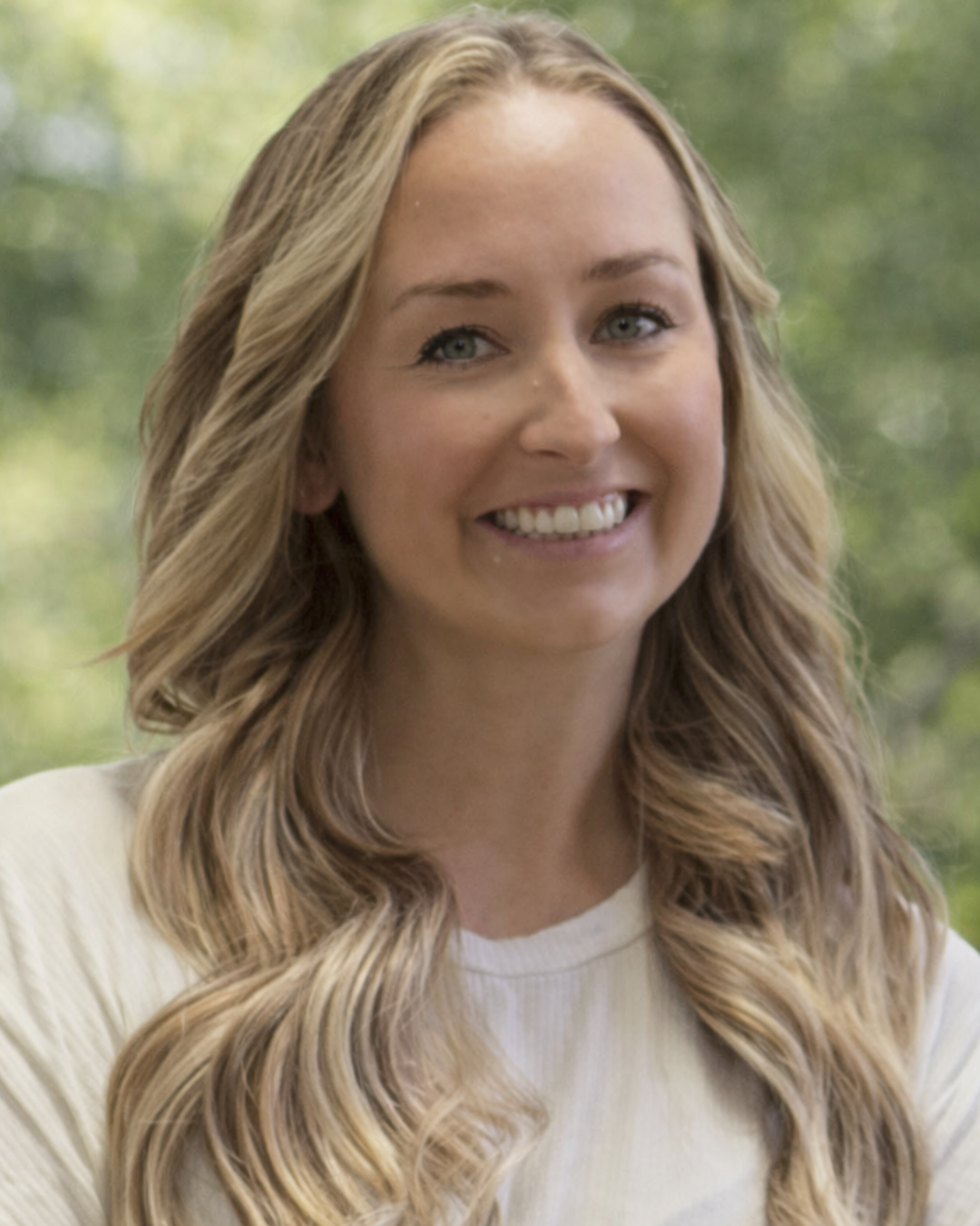 A smiling young woman with wavy blonde hair, wearing a white shirt, standing outdoors with a blurred green background.