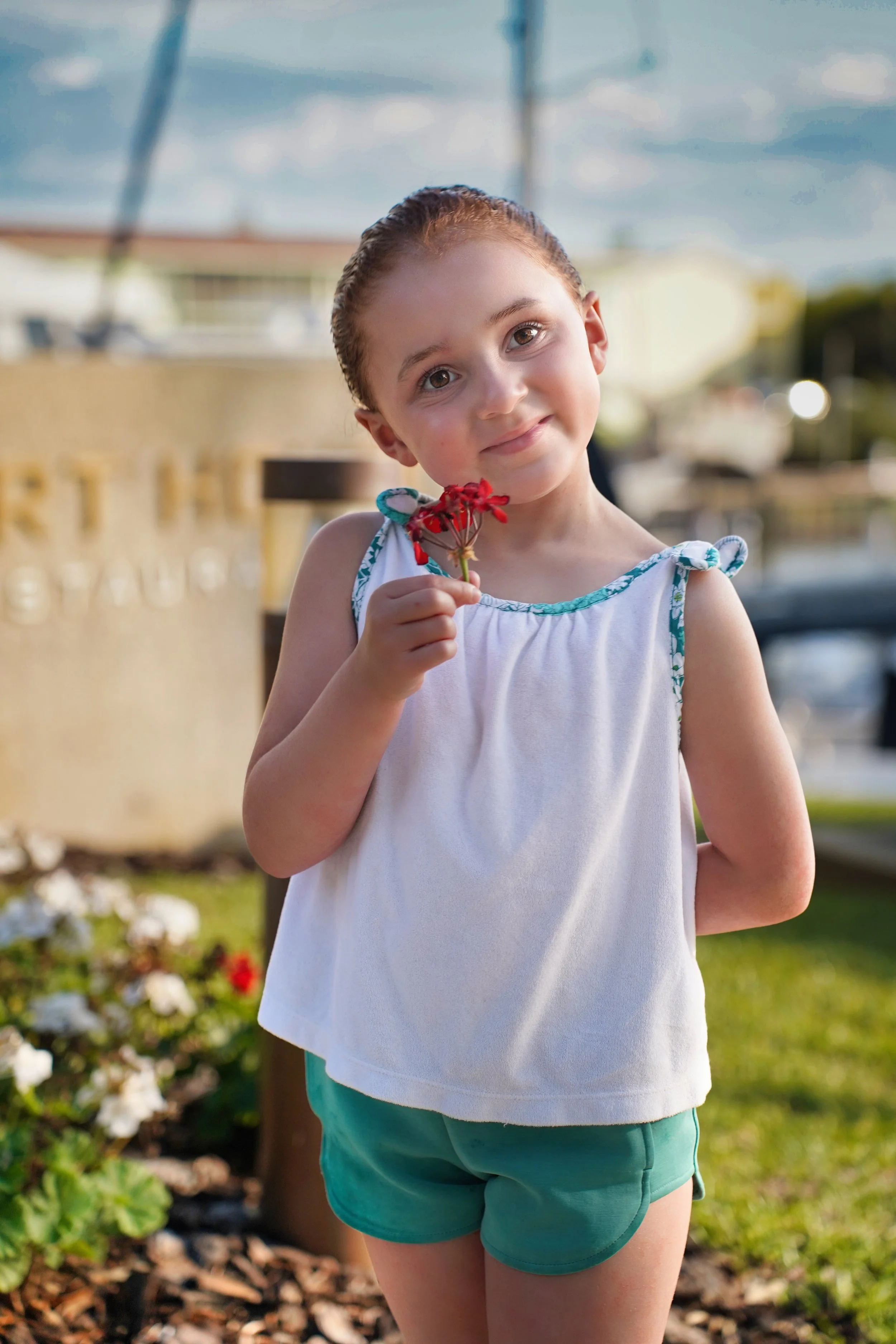A young girl with wet hair holding a red flower, standing outdoors near flowers and greenery.