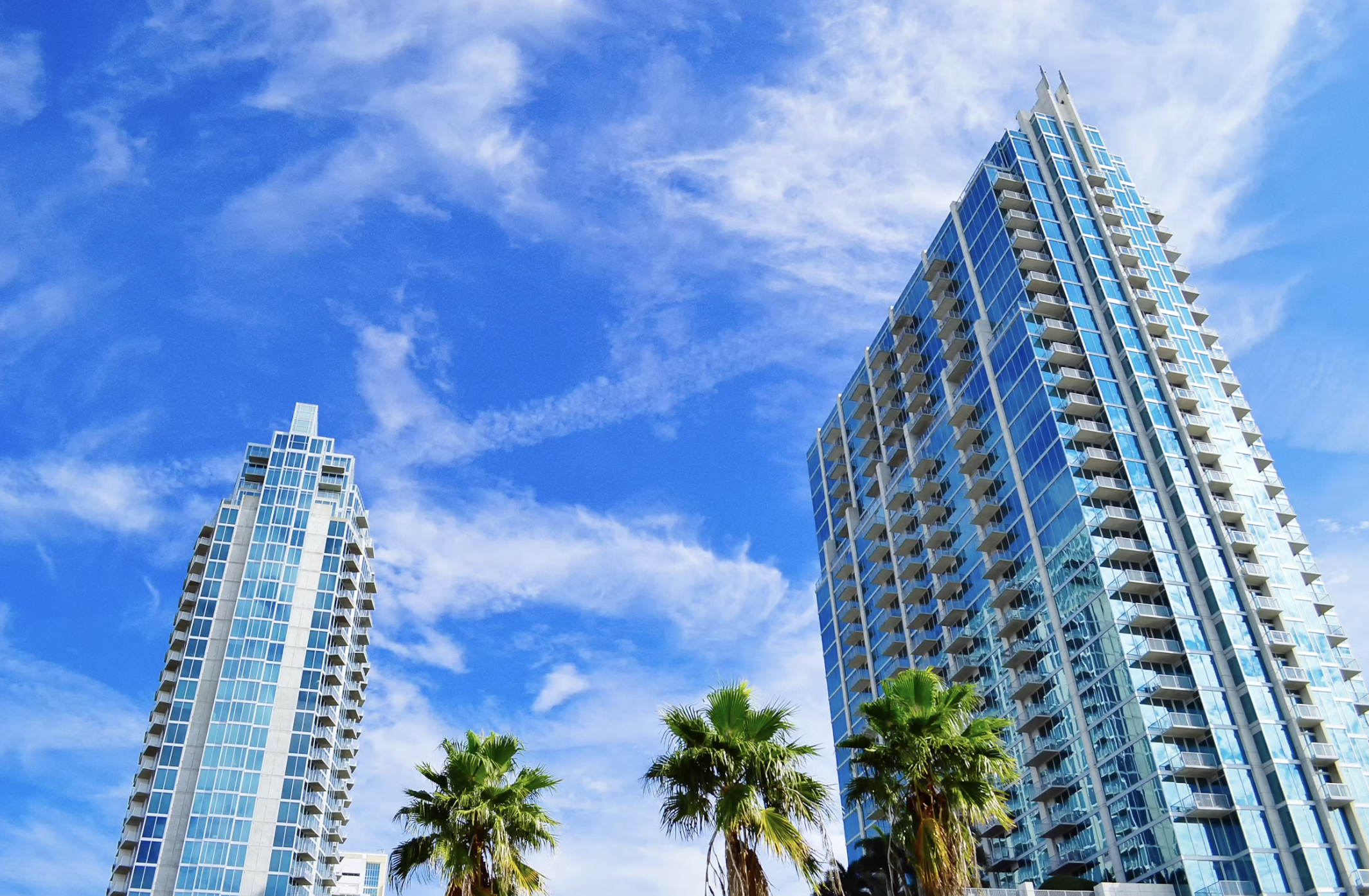 Modern high-rise glass apartment buildings with balconies, under a blue sky with clouds, and palm trees in the foreground.