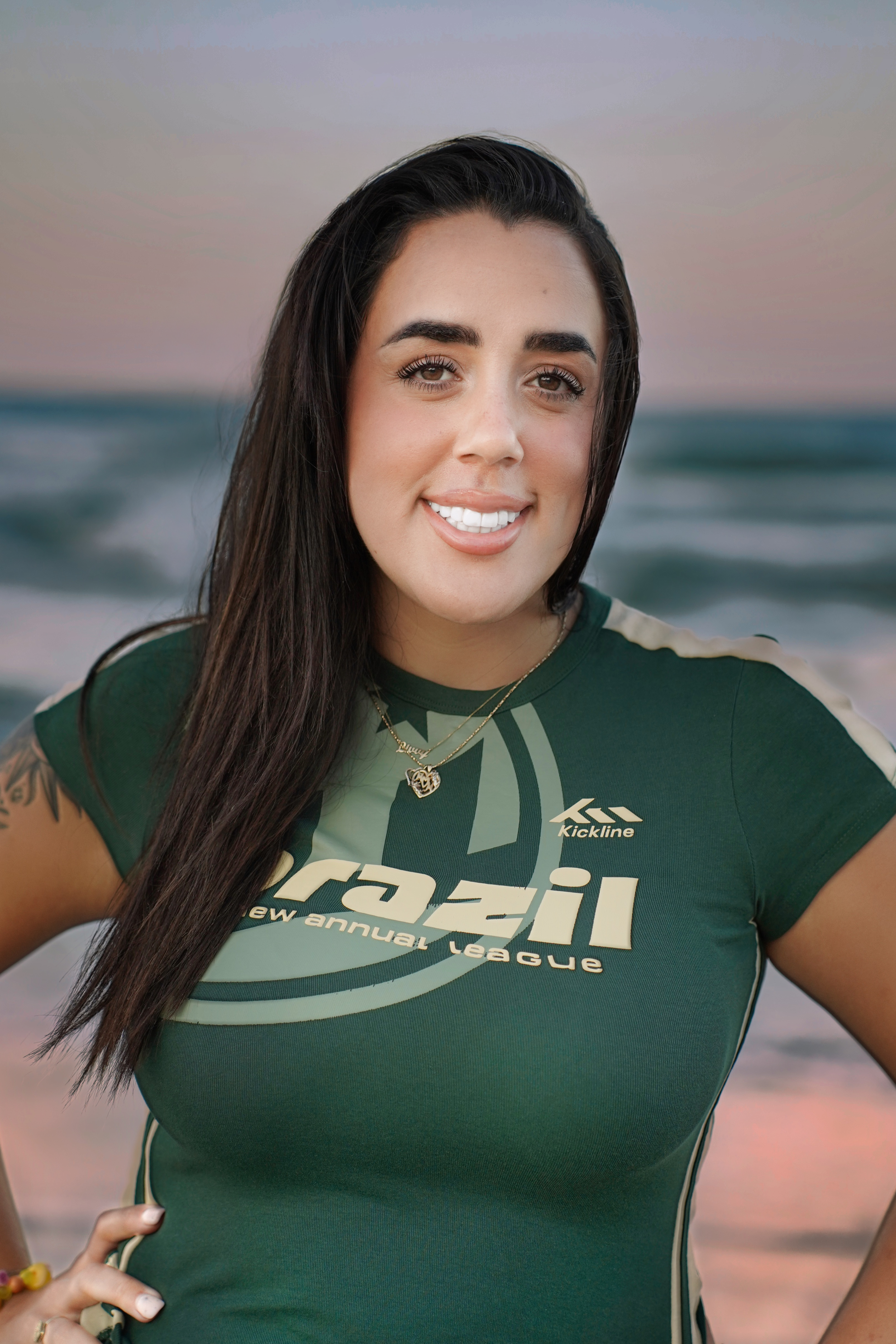 A young woman with long dark hair and light skin smiling at the camera on a beach at sunset, wearing a green athletic shirt with white lettering and jewelry.