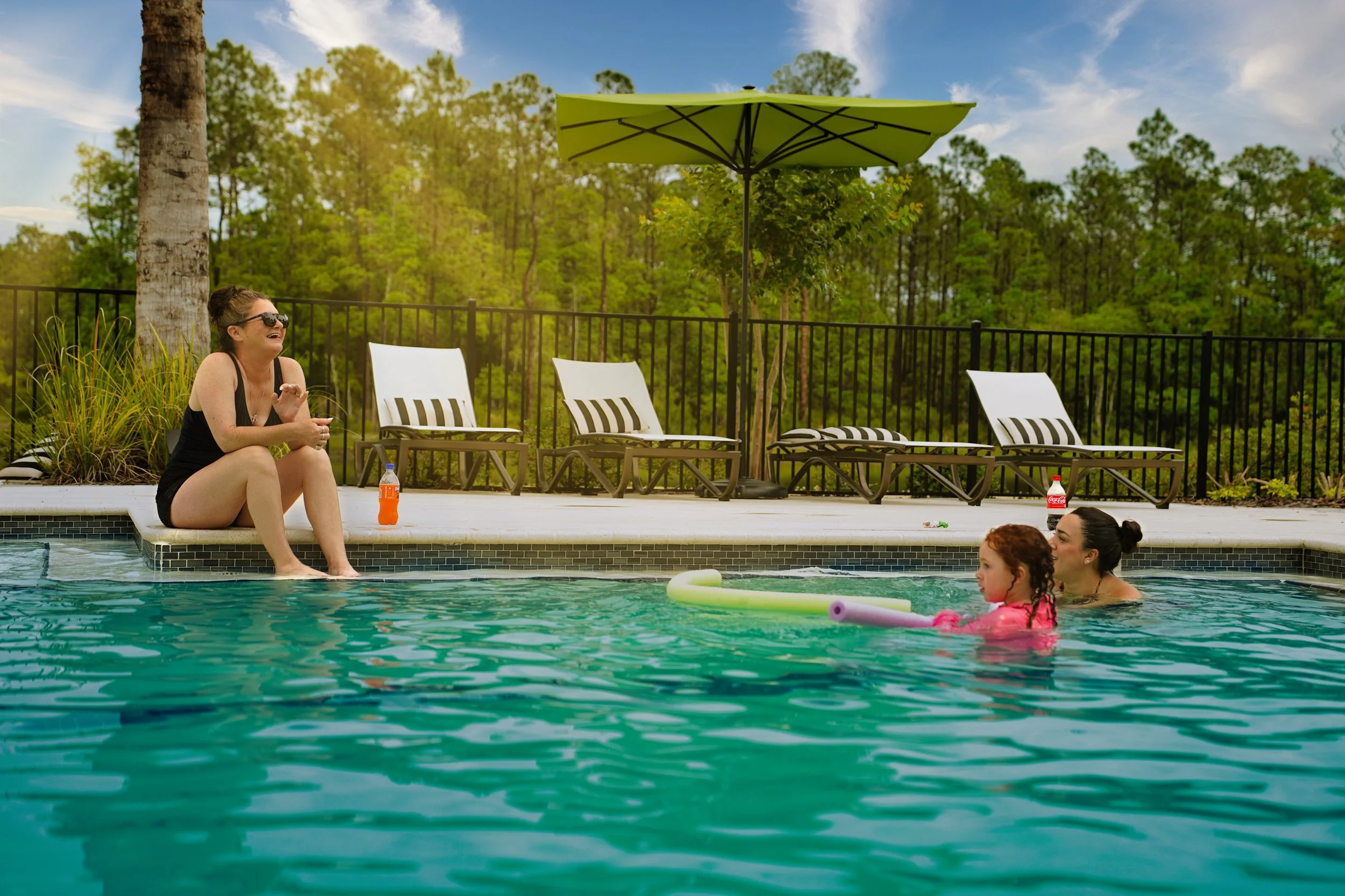 A woman sitting on the edge of a swimming pool Daytona beach, smiling and talking to two children in the pool, with lounge chairs, a large green patio umbrella, and trees in the background. High end photography, the napier
