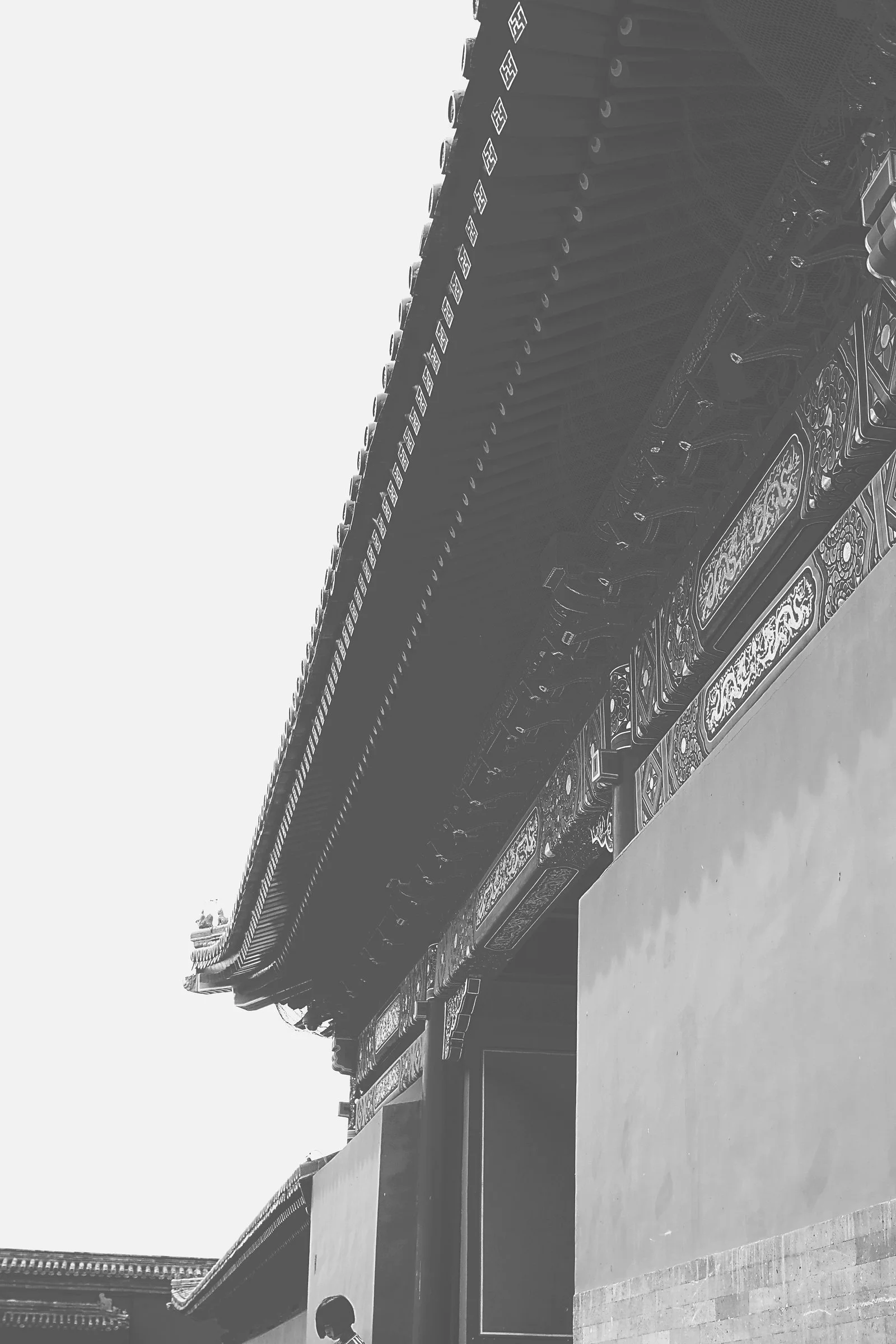 Black and white photo of a traditional Asian temple roof with ornate decorative details, partially obscured by a concrete wall in the foreground, against a clear sky.