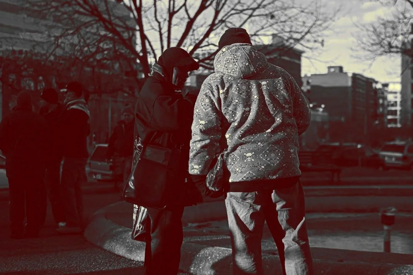 Two men standing on a city sidewalk, engaged in conversation, with other people and buildings in the background. The scene appears to be during late fall or winter.