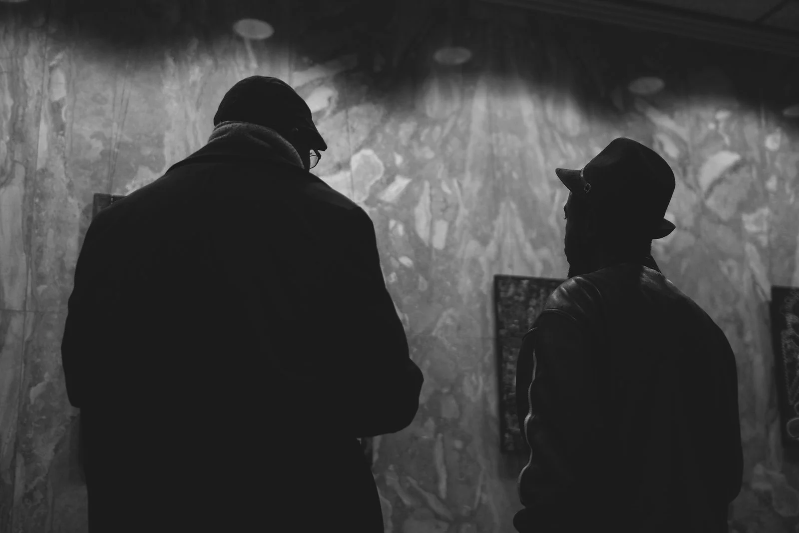 Silhouettes of two people wearing hats, standing in front of a textured wall, engaged in conversation or observation.