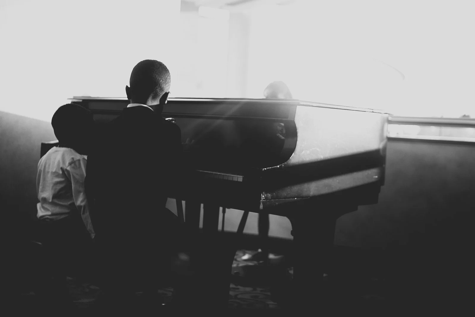 Back view of young boy in a suit playing grand piano in a room with bright light coming through the window.