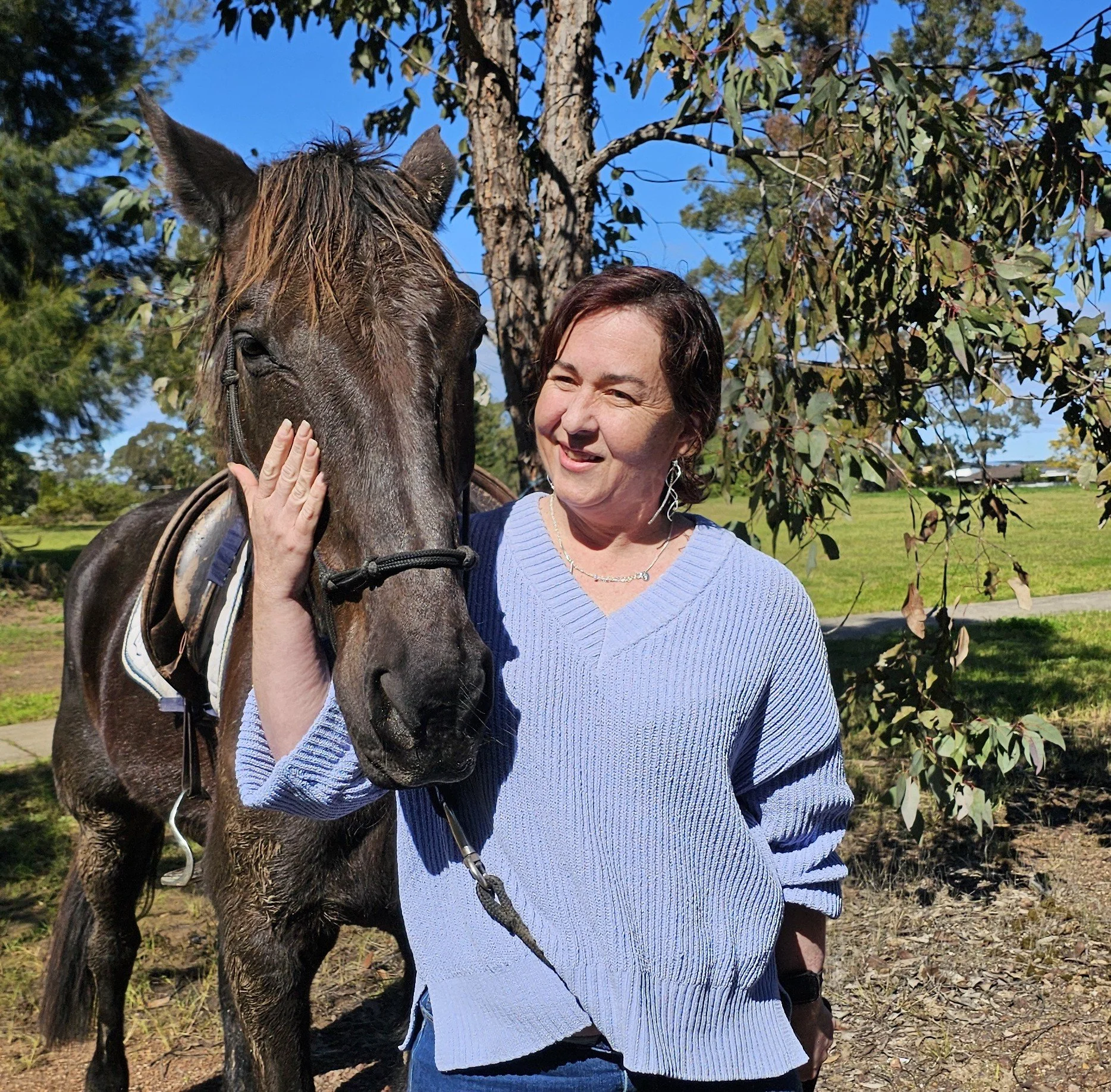 A woman in a lavender sweater smiling while petting a dark brown horse outdoors on a sunny day, with trees and a blue sky in the background.