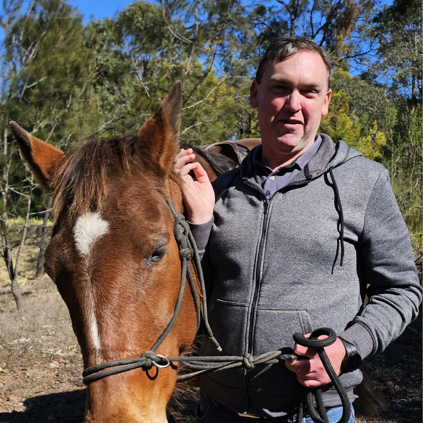 A man with short brown hair wearing a grey zip-up hoodie holds a brown horse's bridle in an outdoor setting with trees and a blue sky in the background.