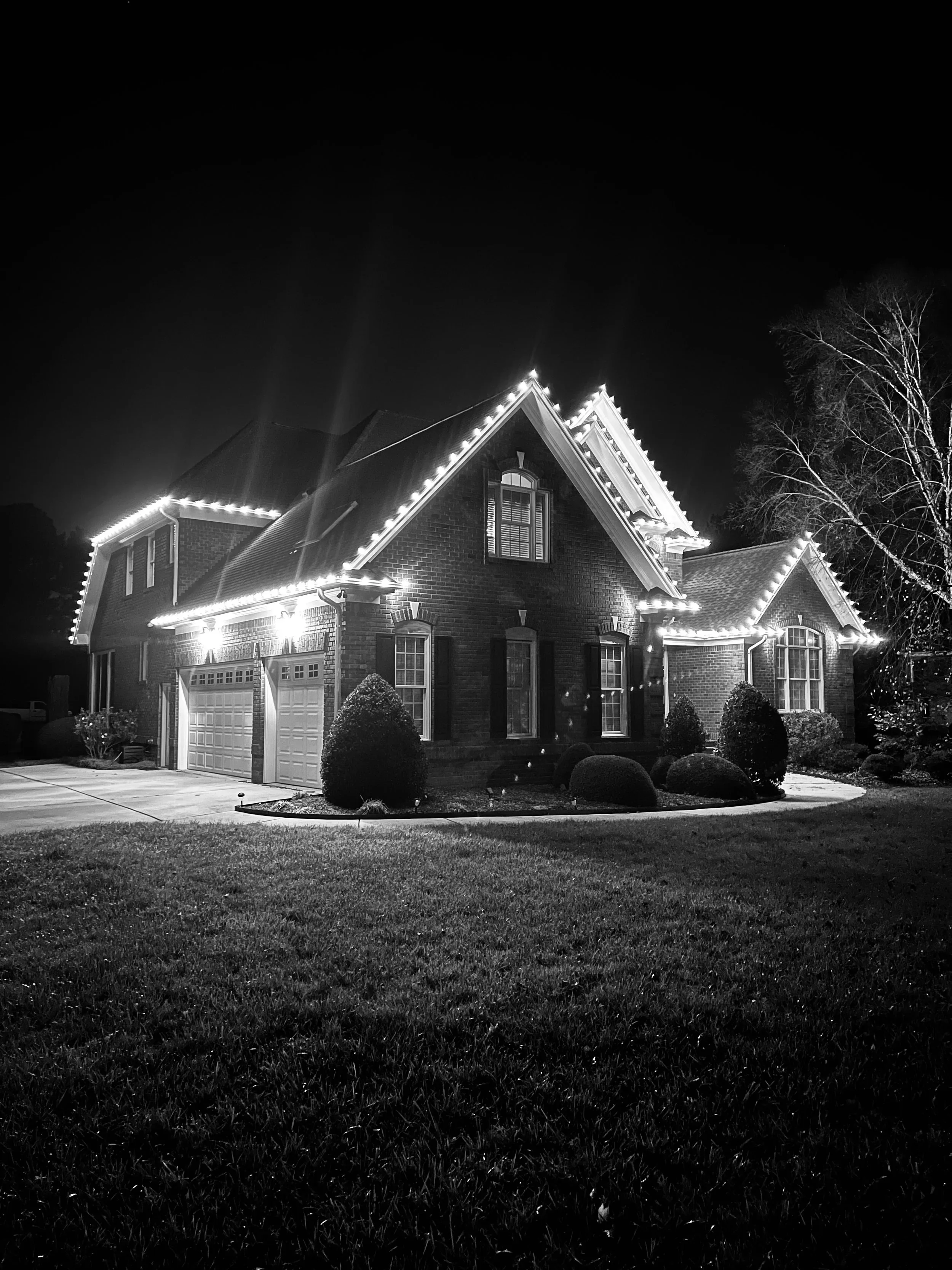 A two-story brick house decorated with Christmas lights at night, with a well-maintained lawn and trees in the yard.