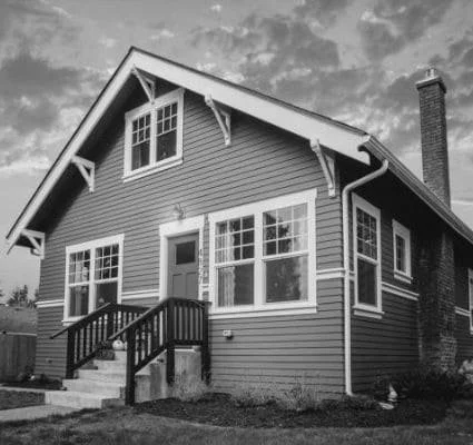 A two-story house with horizontal siding, a front porch with stairs, and multiple windows, under a partly cloudy sky.
