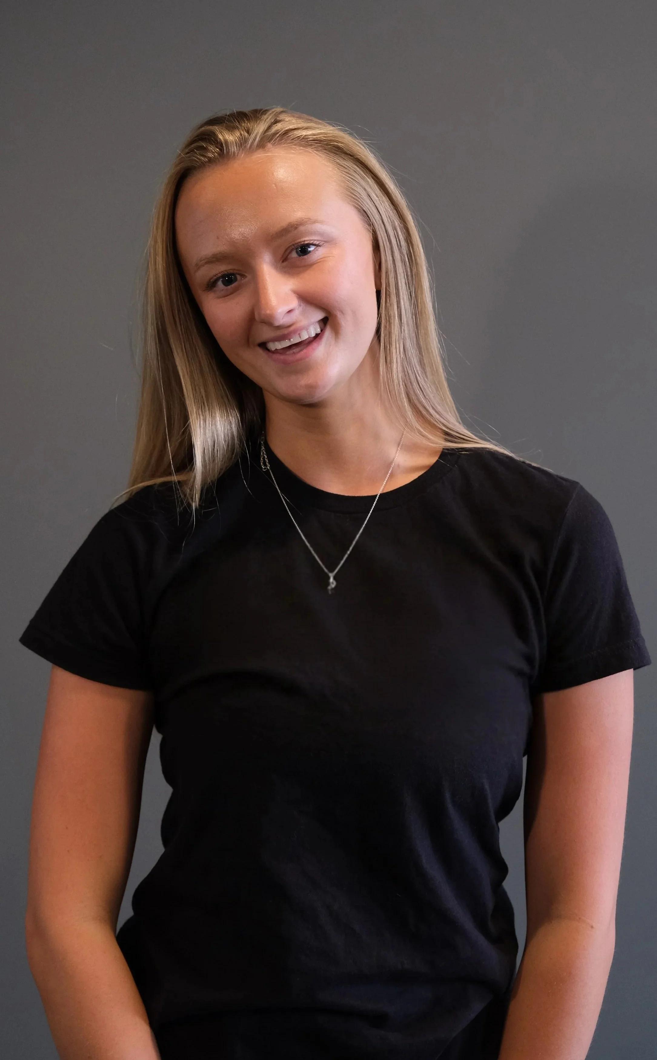 Young woman with long blonde hair wearing a black t-shirt and a silver necklace, standing against a plain dark gray background, smiling slightly at the camera.