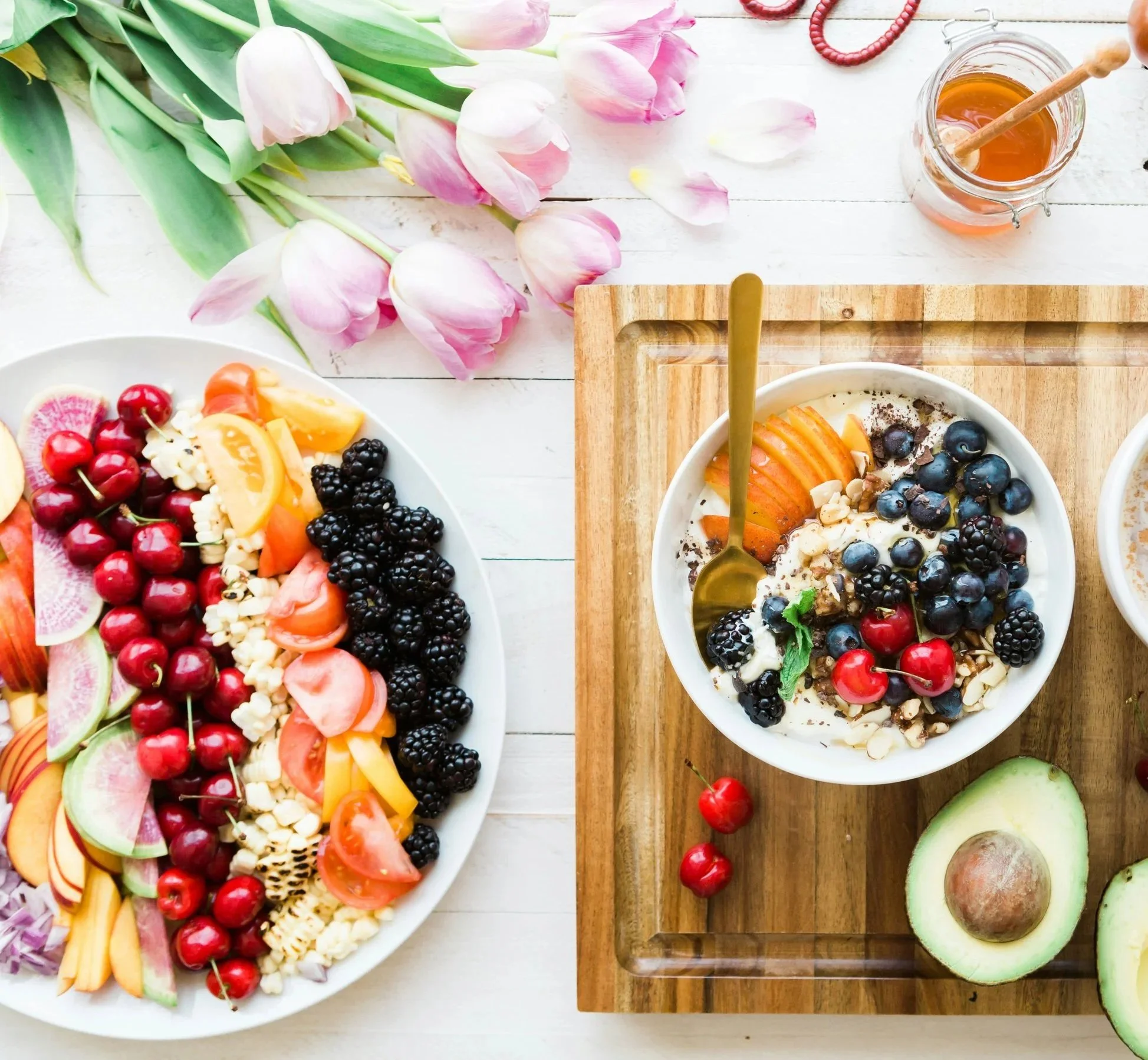 Assorted fresh fruits, granola, and honey on a white table with pink tulips and a wooden tray.