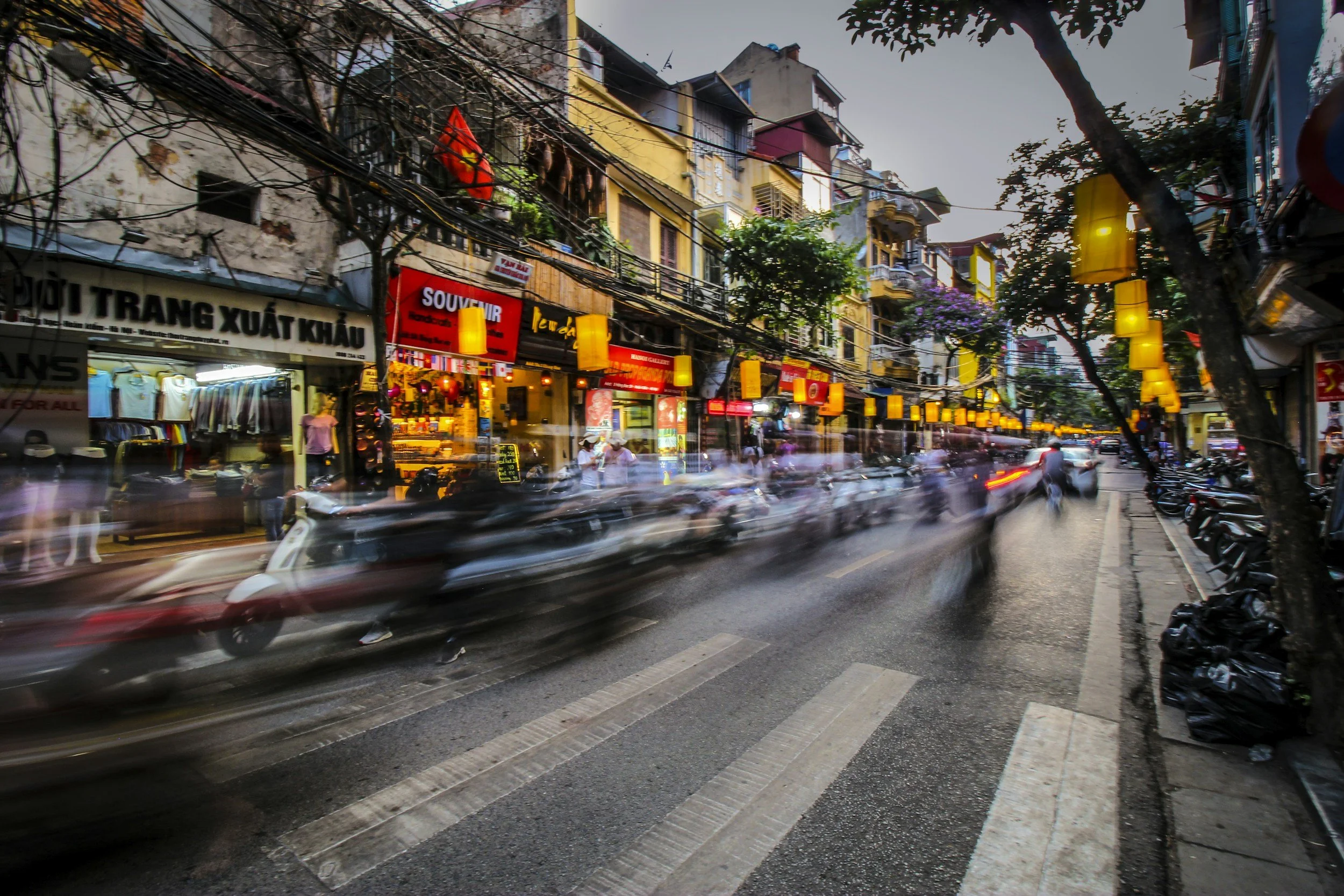 A busy street scene in an urban neighborhood with cars passing by, shops with colorful signs, and multi-story buildings with balconies. Trees line the street, and there are people walking on the sidewalk.