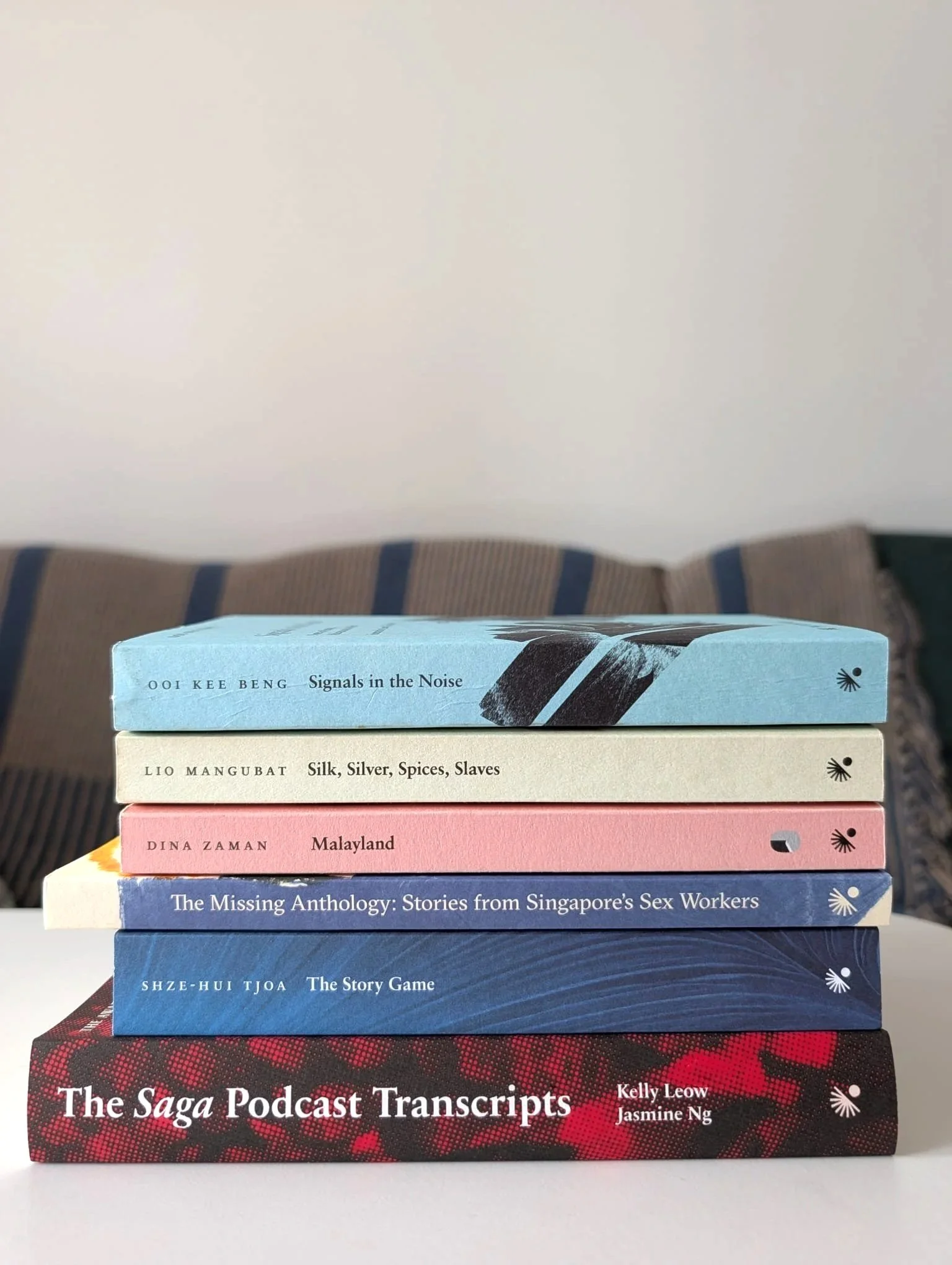 Stacked books with various titles including 'Signals in the Noise', 'Silk, Silver, Spices, Slaves', 'Malayland', 'The Missing Anthology', and 'The Story Game'. The books are on top of a black and red patterned surface, with a plain white background.
