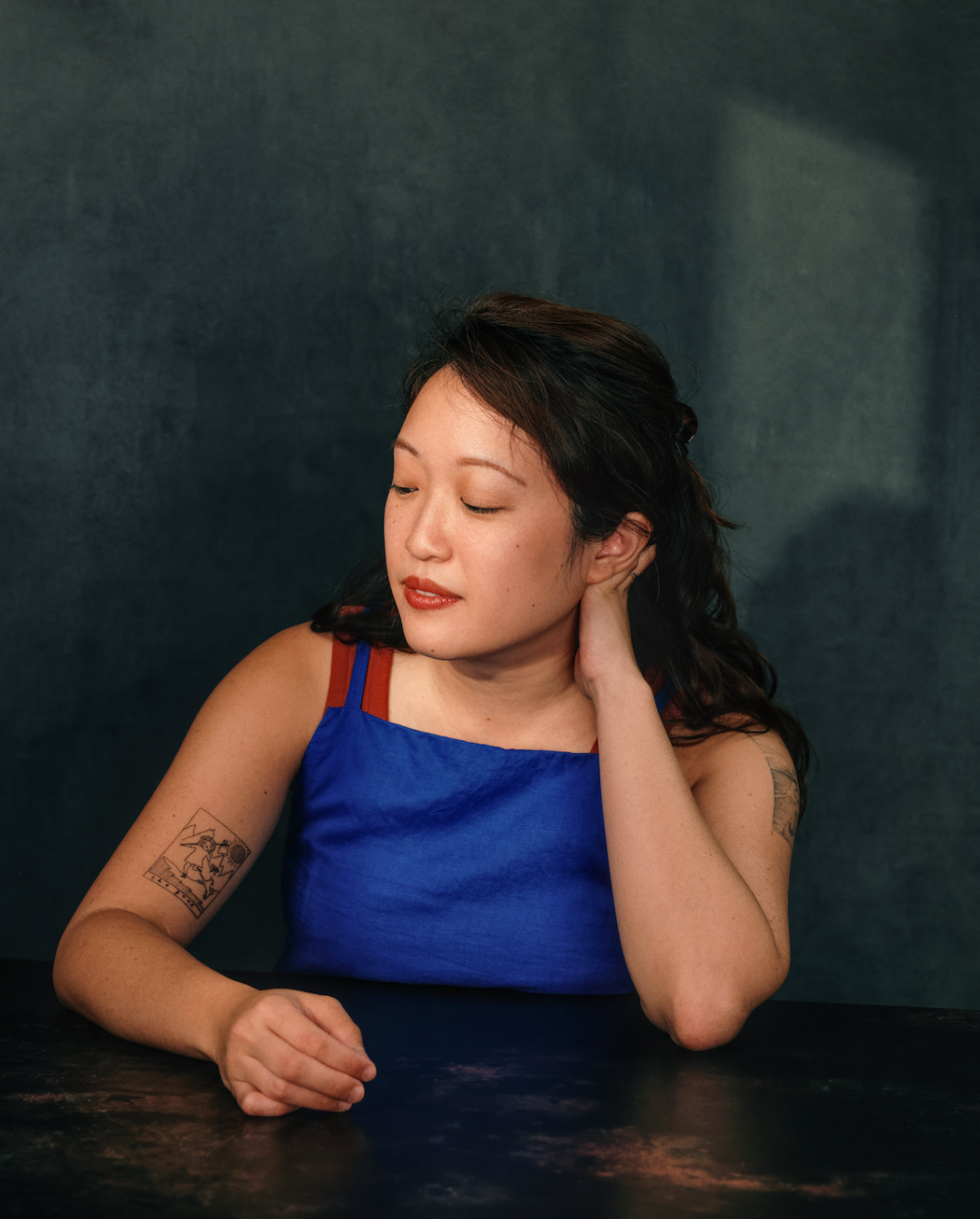 Faction author Shze-Hui Tjoa, wearing a blue top with red straps, sitting at a dark table against a dark background, looking downward with her eyes closed and her hand resting on her neck.
