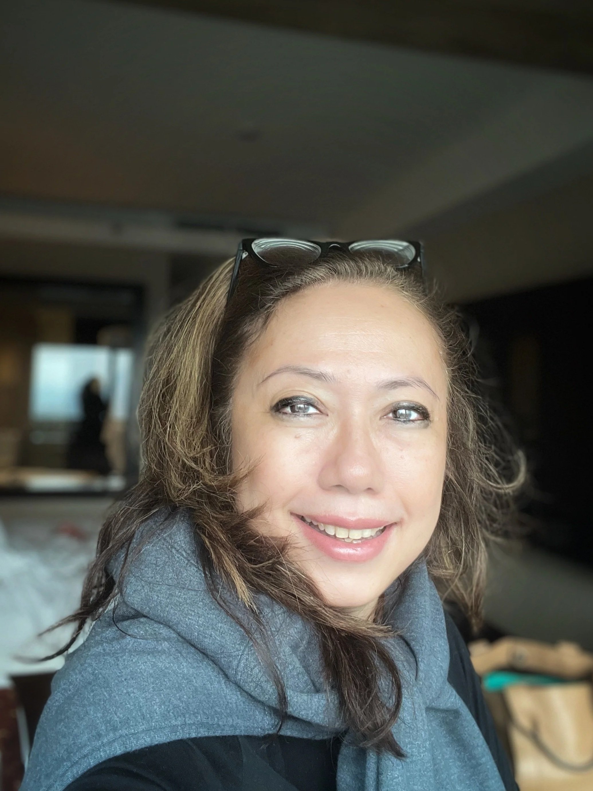 Faction author Dina Zaman pictured here smiling at the camera, wearing glasses on her head and a gray and black jacket, with a blurred background showing an indoor setting.